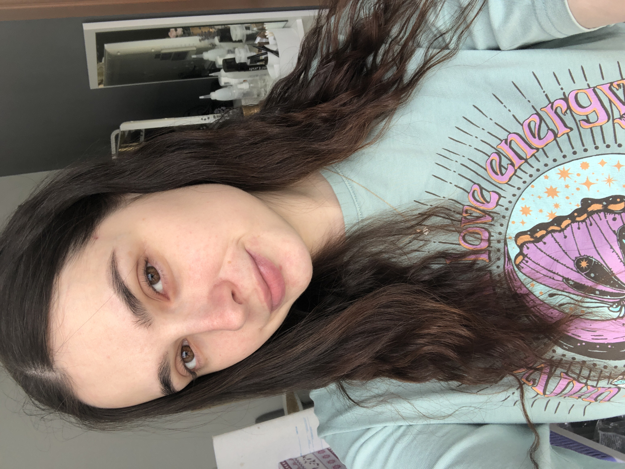 A woman with long, dark brown hair is seen from the chest up in a close-up, slightly angled view. She has fair skin, dark eyebrows, and brown eyes. She wears a light blue t-shirt with a graphic design featuring a butterfly and the word "energy" in a stylized font. In the background, there's a cabinet with items that appear to be cosmetic products. The photo does not provide any information to determine it was taken in Chaikovsky, Russia.