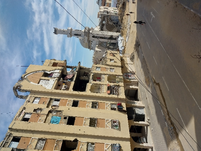 This image depicts a devastated urban street scene in Khan Yunis, Palestinian Territory. In the foreground and left, a multi-story building, likely residential or commercial, exhibits extensive damage, including numerous impact marks consistent with bullet holes or shrapnel, shattered windows, and significant structural collapse on its upper floors, revealing interior spaces and exposed rebar. Some windows are covered with tarps, and clothing hangs from balconies.
To the right and in the midground, a paved road with white lane markings stretches diagonally, featuring a directional arrow pointing forward-right. The road appears uneven in sections, with a darker, wet-looking patch. Four individuals are visible on or adjacent to the road, walking away from the camera. Two are in the mid-distance, one appearing to push or carry an object, while two others are further back.
Beyond the road on the right, a large area of rubble and dirt indicates demolished structures, with white tarps covering some debris piles. In the background, a heavily damaged mosque with a distinct minaret and dome stands amidst the destruction, showing missing sections and visible structural compromise. The sky above is blue with scattered white clouds, and overhead wires traverse the upper portion of the frame.