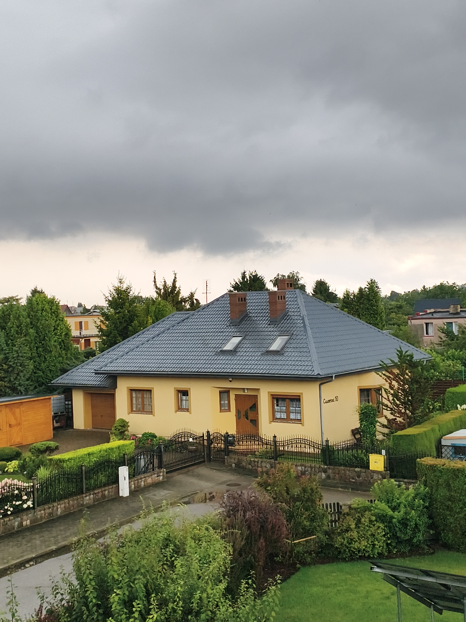 This image depicts a suburban residential area under a cloudy sky, likely in Tczew, Poland. The focal point is a single-story, light yellow house with a dark gray, tiled roof. The house features several windows with wooden frames and a wooden front door. A detached wooden shed is visible to the left of the house. The property is enclosed by a black wrought-iron fence with stone foundations. A paved driveway leads to a garage door on the left side of the house. The yard is landscaped with various green bushes, hedges, and some flowering plants. A white rectangular post stands near the fence. In the background, more houses and mature trees are visible, suggesting a well-established neighborhood. The overall atmosphere is somber due to the overcast weather. There are no people or animals visible in the image.