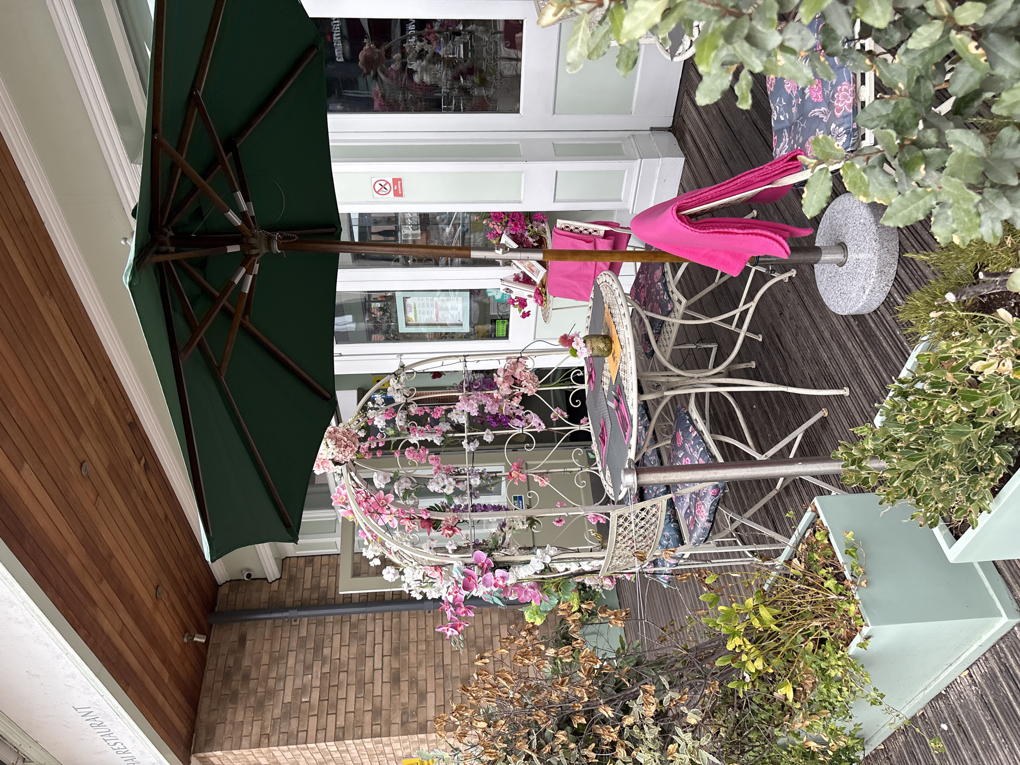This image depicts the outdoor seating area of a Thai restaurant in Chelmsford, United Kingdom. A dark green umbrella shades a white metal table and four chairs, each with floral patterned cushions. The table is set with place settings, and a small vase holds a flower. A decorative white trellis, adorned with artificial pink and white flowers, stands beside the table. To the left of the seating area, a planter box contains dry, brown foliage. The restaurant's facade features large windows and a wooden soffit. A "No Smoking" sign is visible on one of the windows. The seating area is located on a wooden deck.
