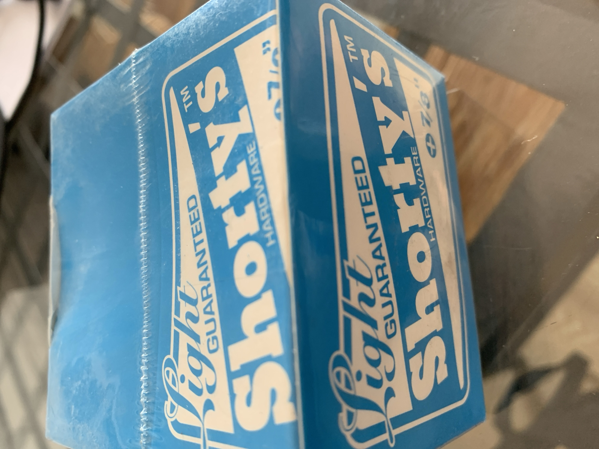 The image shows a close-up of a blue box with white lettering, likely containing hardware. The box is wrapped in clear plastic. The lettering on the box appears to be "Shorty's" and "Light Guaranteed". The bottom portion of the box is partially obscured by a reflective surface, possibly a table. The background is blurred and appears to be an indoor setting. The image does not contain any information that would allow for determining its location in Barcelona, Spain.