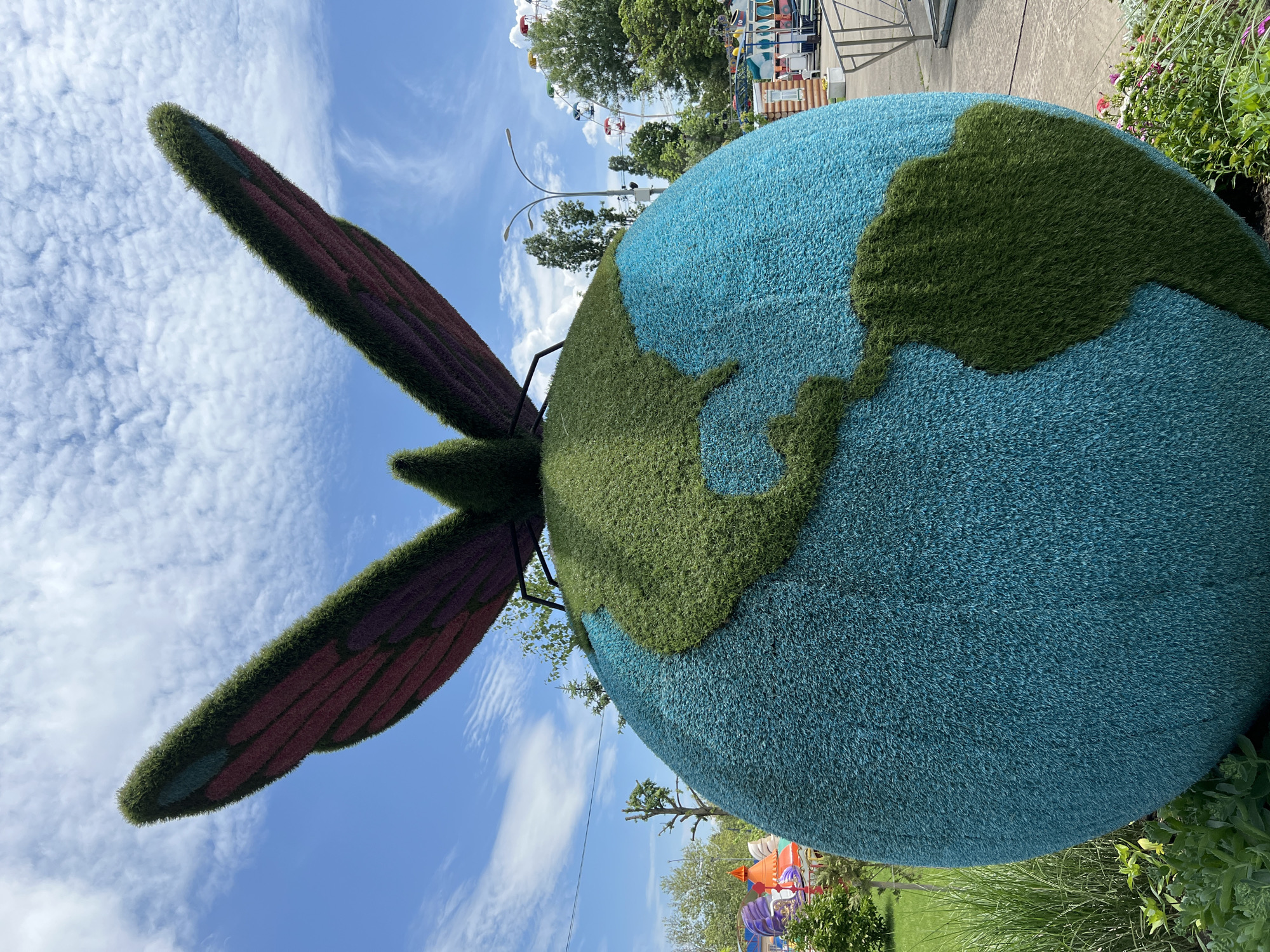 The image shows a large, spherical art installation resembling a globe, with continents depicted in green and oceans in blue. A butterfly with green, purple, and red wings appears to be affixed to the globe. The installation is situated outdoors, possibly in a park or public space, with a blue sky and scattered clouds in the background. A Ferris wheel is visible in the distance, along with other amusement park rides. Some greenery is visible at the base of the globe and along the edges of the frame. The location is Krasnodar, Russia.