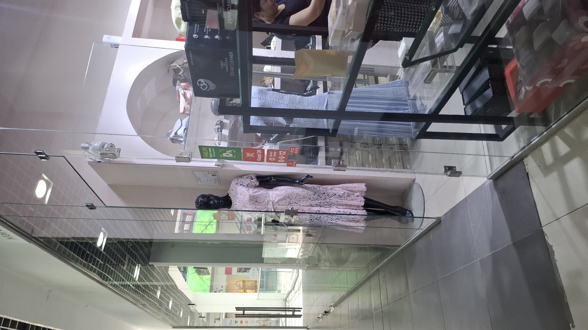 A shop window in a mall in Shymkent, Kazakhstan, features a black mannequin dressed in a pink lace dress. Behind the mannequin, on shelves, are various items including handbags and boxes, some of which are labeled "GERMAN QUALITY" and "STEAM CLEANER." Another mannequin, this one dressed in a light blue dress, is visible further inside the shop. A woman is partially visible in the background, attending to a display of items. Signs are posted on the glass, one with a "0%" graphic and another for "Kaspi QR." The floor is tiled, and the shop entrance is framed by glass panels and a metal door handle. The environment appears to be a retail space within an indoor shopping center.