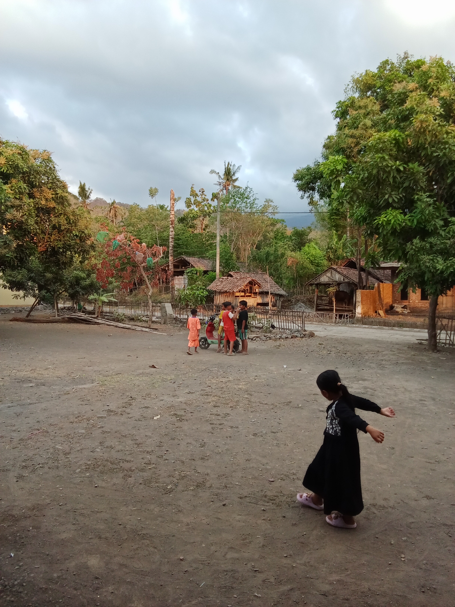 The image depicts a rural scene in Selowogo, Indonesia, characterized by a dusty, open area in the foreground and a village settlement in the background. The sky is overcast, with a muted, twilight-like light. In the foreground, a young girl in a black dress and pink sandals is captured mid-motion, with her arms extended, appearing to be dancing or playing. Further back, a group of three children, two boys and a girl, are gathered around a small red scooter. They are dressed in casual, brightly colored clothing. Behind the children, traditional Indonesian houses with thatched or corrugated metal roofs are visible, nestled amongst lush greenery and trees. The environment suggests a warm climate, with the vegetation indicating a tropical or subtropical setting. The overall impression is one of everyday village life in a developing region.