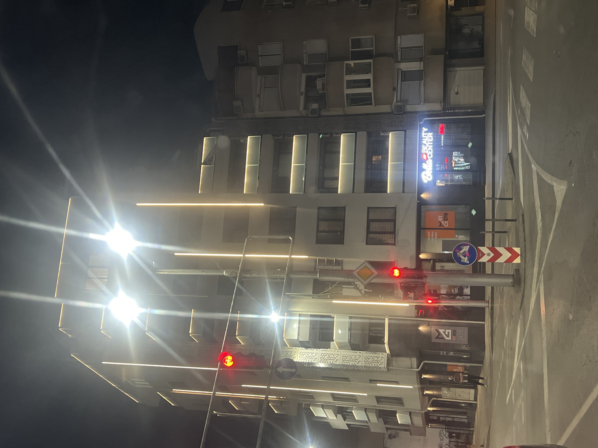 The image shows an urban scene at night, likely in Plovdiv, Bulgaria, given the signage in Cyrillic. The street is illuminated by streetlights, and buildings are visible on both sides. A modern building with vertical light strips is prominent on one side, and a sign that says "Bella Beauty Center" is visible on one of the buildings. There is a traffic light showing a red light, along with various road signs. There is at least one pedestrian in the crosswalk. The street appears to be wet, possibly from recent rain. The buildings are multi-storied, with some having air conditioning units. The general impression is a nighttime city scene, with functional buildings and traffic management.