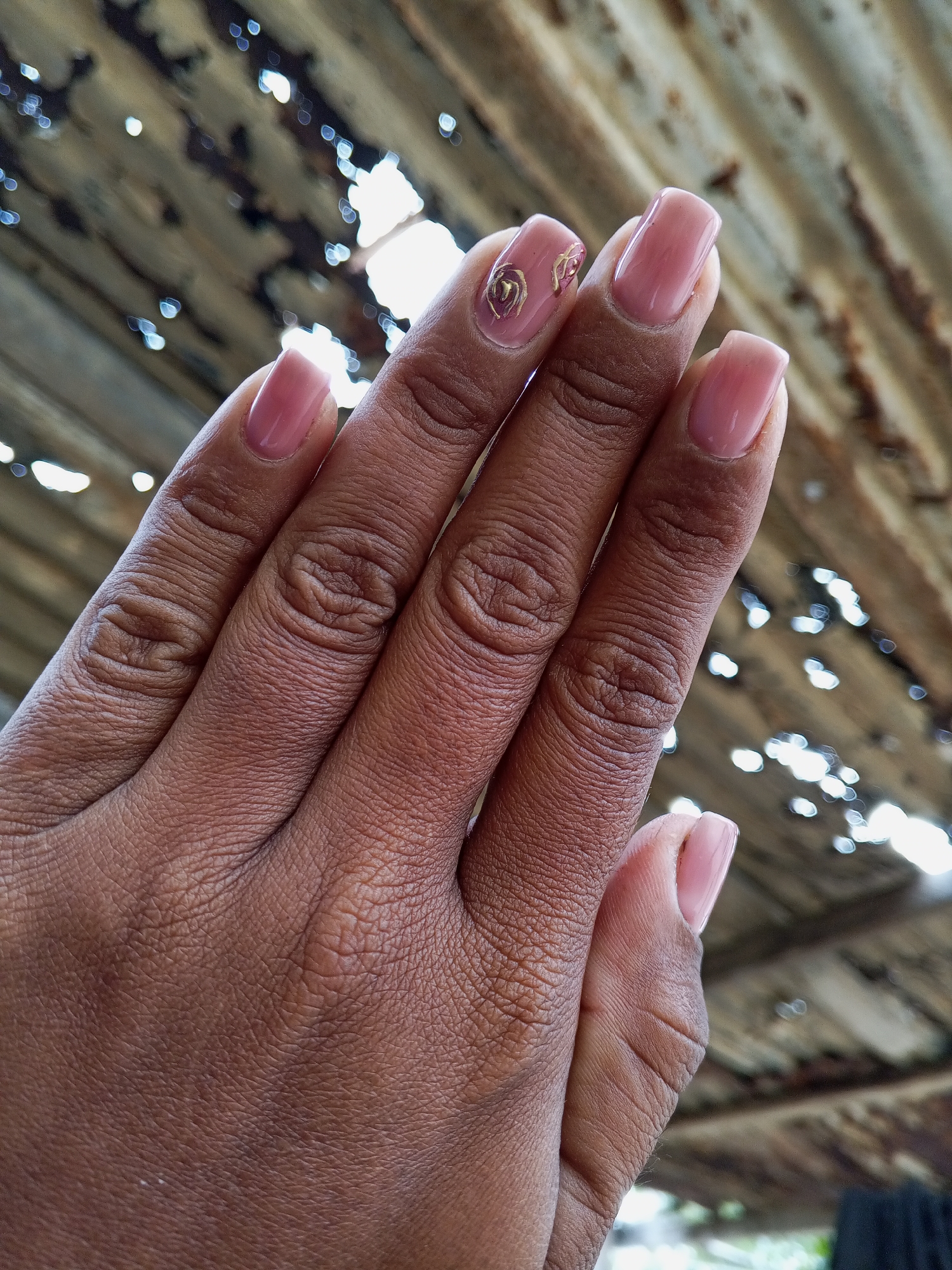 The image presents a close-up view of a person's left hand, angled upwards and slightly open, prominently displaying a fresh manicure against a heavily blurred background. The hand has a dark brown skin tone, showing detailed skin texture and creases on the knuckles.
Each fingernail is neatly manicured into a rounded square shape and coated with a glossy, translucent light pink or nude-colored polish. A distinctive decorative element is visible on the ring finger's nail: a delicate, metallic gold-colored design, possibly an abstract swirl or a stylized heart.
The background is deeply out of focus, characterized by numerous bright, circular bokeh lights. The underlying structure of the background appears corrugated, suggesting an old metal roof or similar weathered material, viewed from an angle that allows light to filter through or reflect brightly. The bright, glinting lights indicate the photograph was taken during daylight hours, likely outdoors or in a semi-outdoor setting with ample natural light.
No specific activity is taking place beyond the pose showcasing the manicured nails. There are no discernible emotions conveyed, and no visible text is present in the image. The setting appears informal and somewhat rustic due to the background's texture and condition.