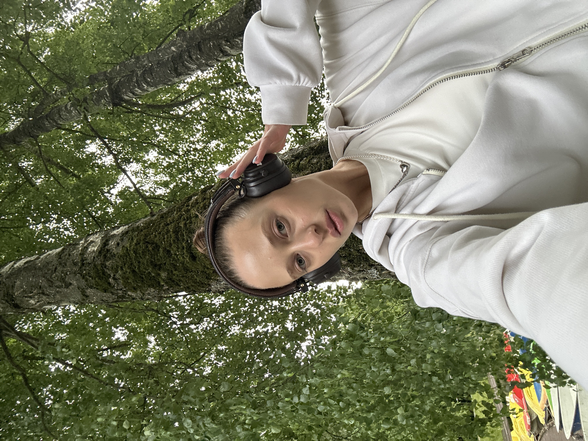 The image shows a young woman wearing a white hoodie and black headphones, holding one hand to her head. She is outdoors, with a large tree trunk and green foliage in the background. The lighting suggests it is daytime, likely overcast or in a shaded area due to the soft light. The woman has light skin, light brown hair pulled back, and is looking directly at the camera with a neutral expression. Her headphones appear to be over-ear style. Part of a zipped hoodie and a silver necklace are visible. In the background, colorful banners or flags can be seen amongst the trees, hinting at a festive or outdoor event. The presence of lush greenery and a large tree suggests a natural setting, possibly a park or forest. The overall mood is casual and relaxed. The location context is Krasnogorsk, Russia.