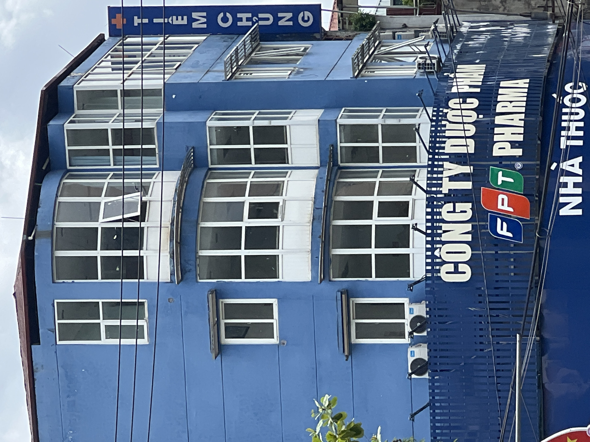 The image shows a blue, multi-story building with a series of windows. A blue sign with white text is attached to the right side of the building. The sign reads "TIEM CHUNG" in vertical orientation. Below the building, a blue fence is visible with white lettering that says "CONG TY DUOC PHAM" and "FPT PHARMA". Another blue sign with white lettering below this reads "NHA THUOC". There are also two air conditioning units mounted on the exterior of the building above the fence. Power lines are strung across the scene. The sky is overcast. The location is specified as Hà Giang 2 Ward, Vietnam.