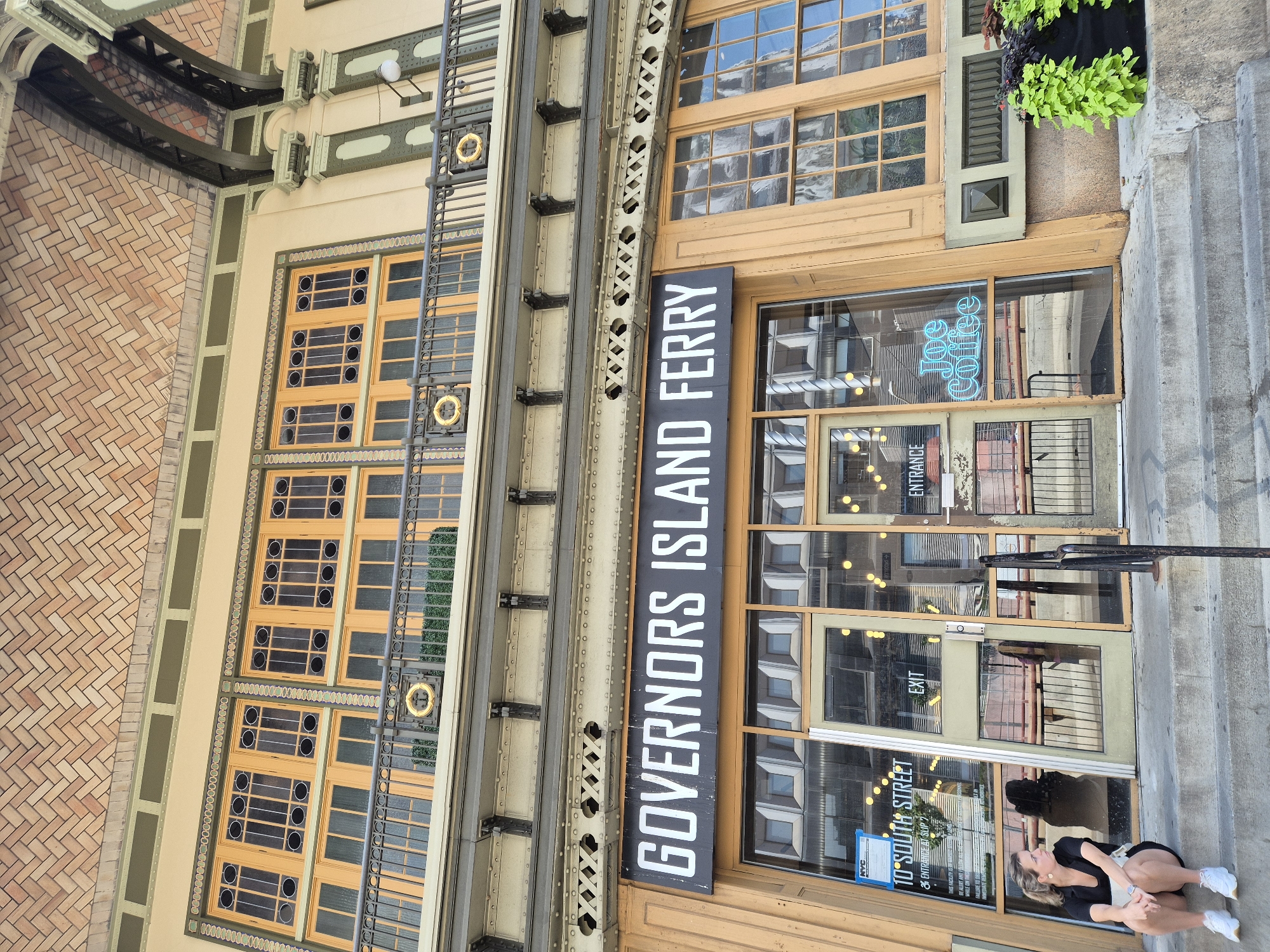 This is a ground-level, eye-level, outdoor, full-shot image of the entrance to the Governors Island Ferry in New York City. The building features a pale yellow facade with dark green trim and large, multi-paned windows. Above the entrance, a prominent black sign reads "GOVERNORS ISLAND FERRY" in white block letters. The ferry terminal is located at 10 South Street.
To the left of the entrance, a young woman with blonde hair tied back is sitting on the concrete steps, facing left. She is wearing a black short-sleeved shirt, black shorts, and white sneakers. She is looking off to the left of the frame and holding a light-colored object in her lap.
The glass doors of the entrance are visible, with signs for "EXIT" and "ENTRANCE." Through the glass, a neon blue sign spelling "Joe Coffee" is illuminated. Inside the building, people and interior lighting are reflected in the windows. Planters with vibrant green foliage are situated to the right of the entrance. The architecture suggests an older, possibly historical building with decorative elements on the upper floors, including arched windows and a tiled roof.