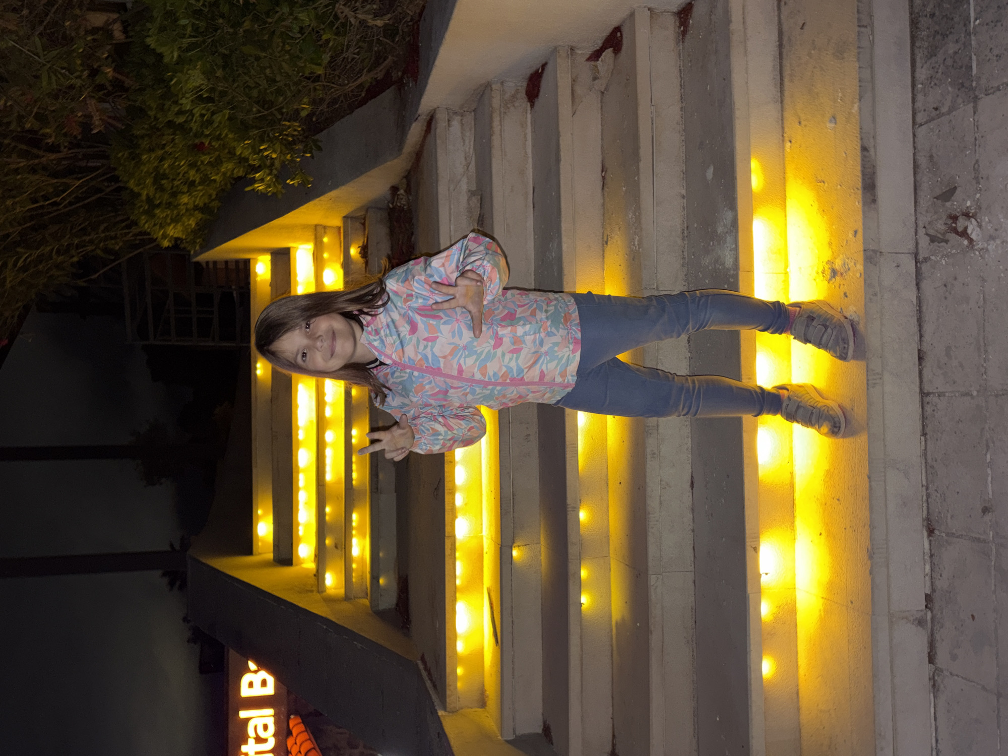 A young girl stands on a set of stairs illuminated with yellow lights in Sveti Stefan, Montenegro. She is wearing a patterned jacket and jeans. The stairs appear to be part of an outdoor structure, possibly leading to a building or garden, with greenery visible in the background. The scene is captured in low light, likely at dusk or night. The girl is making a hand gesture.