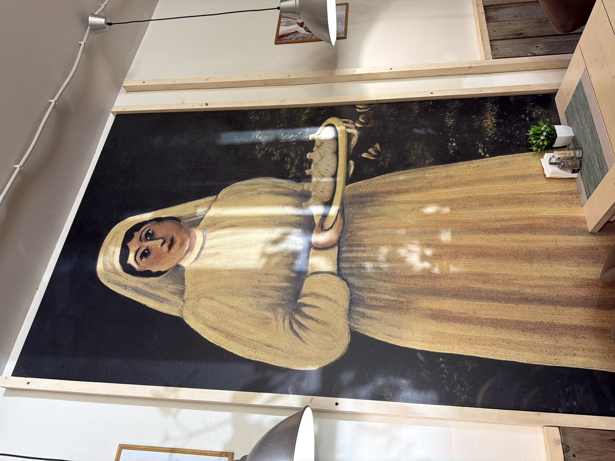 The image shows a large portrait of a nun in a yellow habit and black veil hanging on a wall. The background of the portrait is a dark forest. The portrait is framed with light-colored wood. Above the portrait, there is a picture in a gold frame. There is a light fixture shining directly on the portrait. A table with a green placemat, a small plant, and condiments is visible to the right of the painting. The location is not explicitly stated as Moscow, Russia.