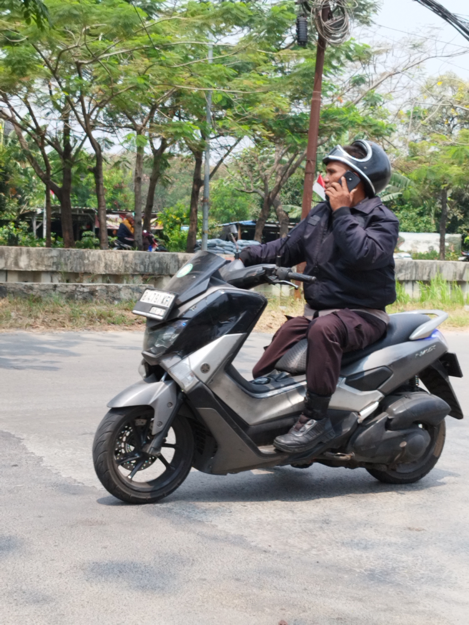 A man wearing a dark jacket, dark pants, and a black helmet is riding a silver and black scooter on a paved road. He is holding a black smartphone to his right ear with his left hand and appears to be speaking on it. The scooter has a license plate that reads "B 7931 KJH". Behind the scooter, there is a concrete wall, followed by a line of trees with green foliage. The scene appears to be outdoors, possibly in an urban or suburban area in Bekasi, Indonesia, given the presence of common Indonesian elements like the scooter and the style of vegetation. The road is asphalt, with some debris and shadows on its surface. The sky is bright and appears to be daytime.