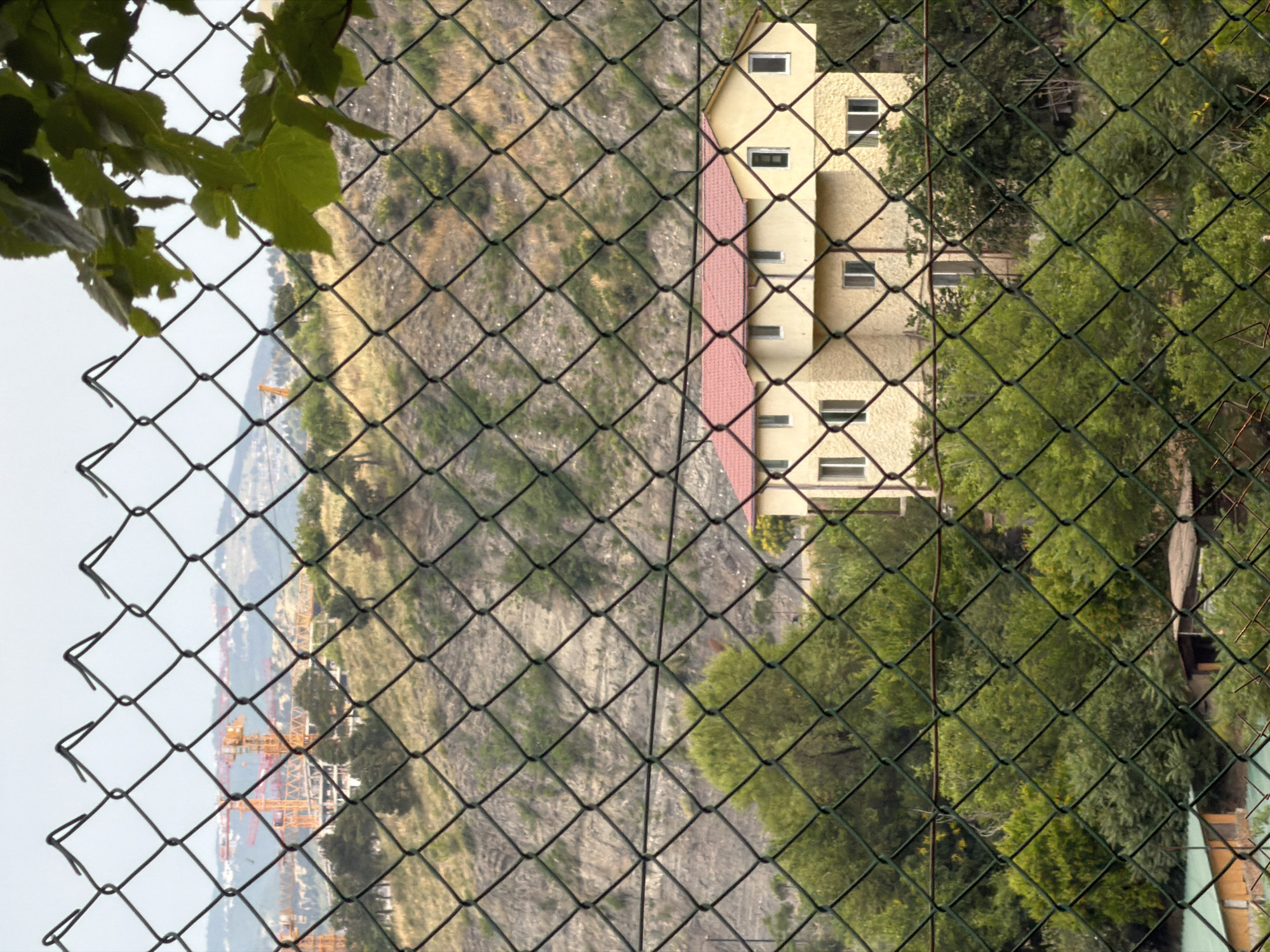 An outdoor, medium-distance, eye-level, vertical shot captures a light yellow, two-story house with a red tiled roof, situated amidst lush green trees. The house features multiple rectangular windows and is partially obscured by a dark green chain-link fence in the foreground. In the background, a steep, barren hillside with patches of dry grass and scattered greenery rises, leading to a hazy skyline. Further in the distance, construction cranes are visible, indicating ongoing development. The scene appears to be located in Tbilisi, Georgia, given the characteristic hilly terrain and construction activity. The lighting suggests an overcast day.