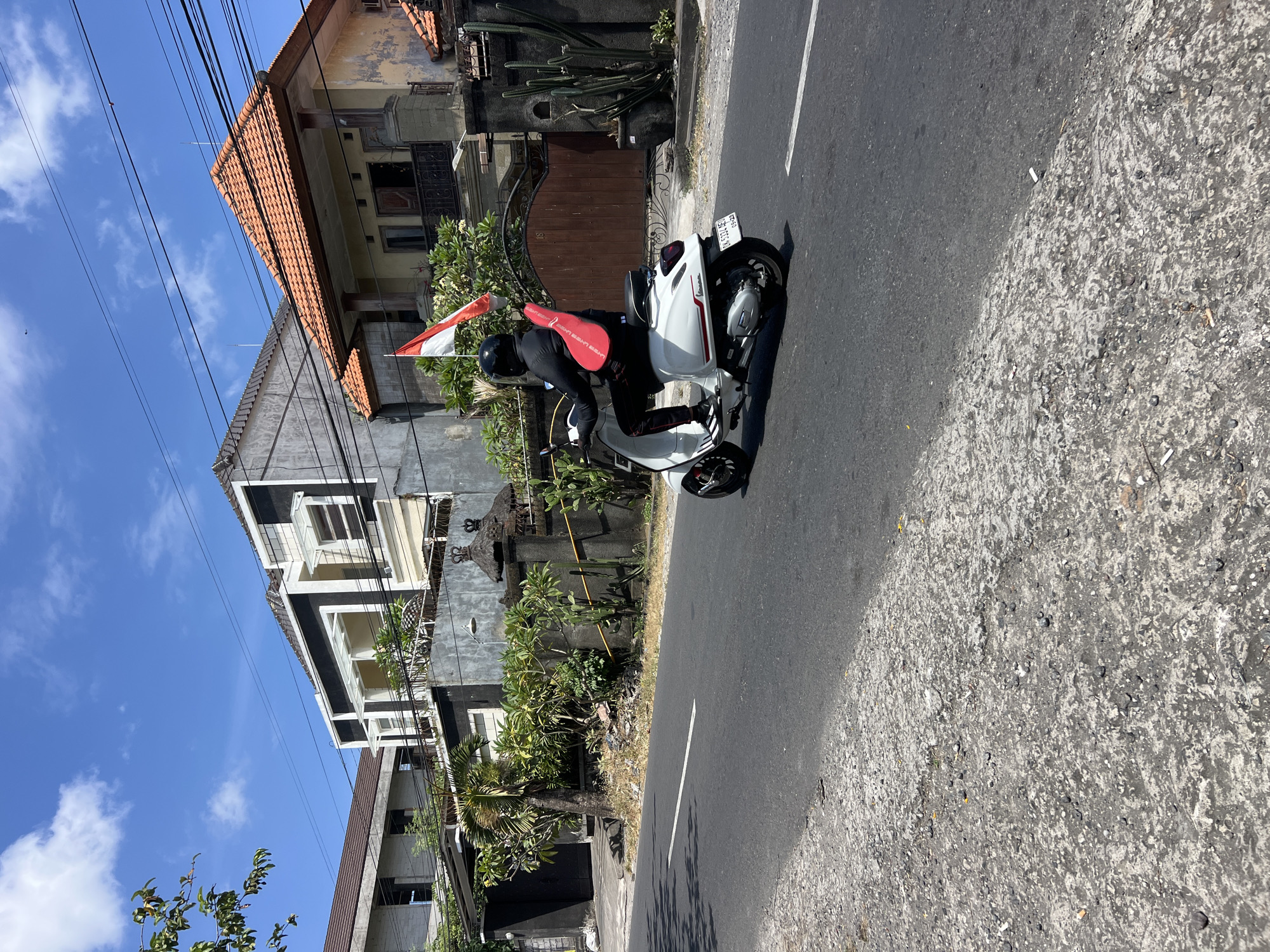 A person wearing a black helmet and black clothing rides a white Vespa scooter down a paved road in Denpasar, Indonesia. A red bag is slung over the rider's back, and an Indonesian flag is mounted to the rear of the scooter. The scooter has a license plate that reads "DK 5371 AE." The road is bordered by a rough, grey shoulder with scattered debris. In the background, there are several multi-story houses with various architectural styles, lush green vegetation, a large wooden gate, and a bright blue sky with scattered clouds. The scene appears to be set in a residential area on a sunny day.