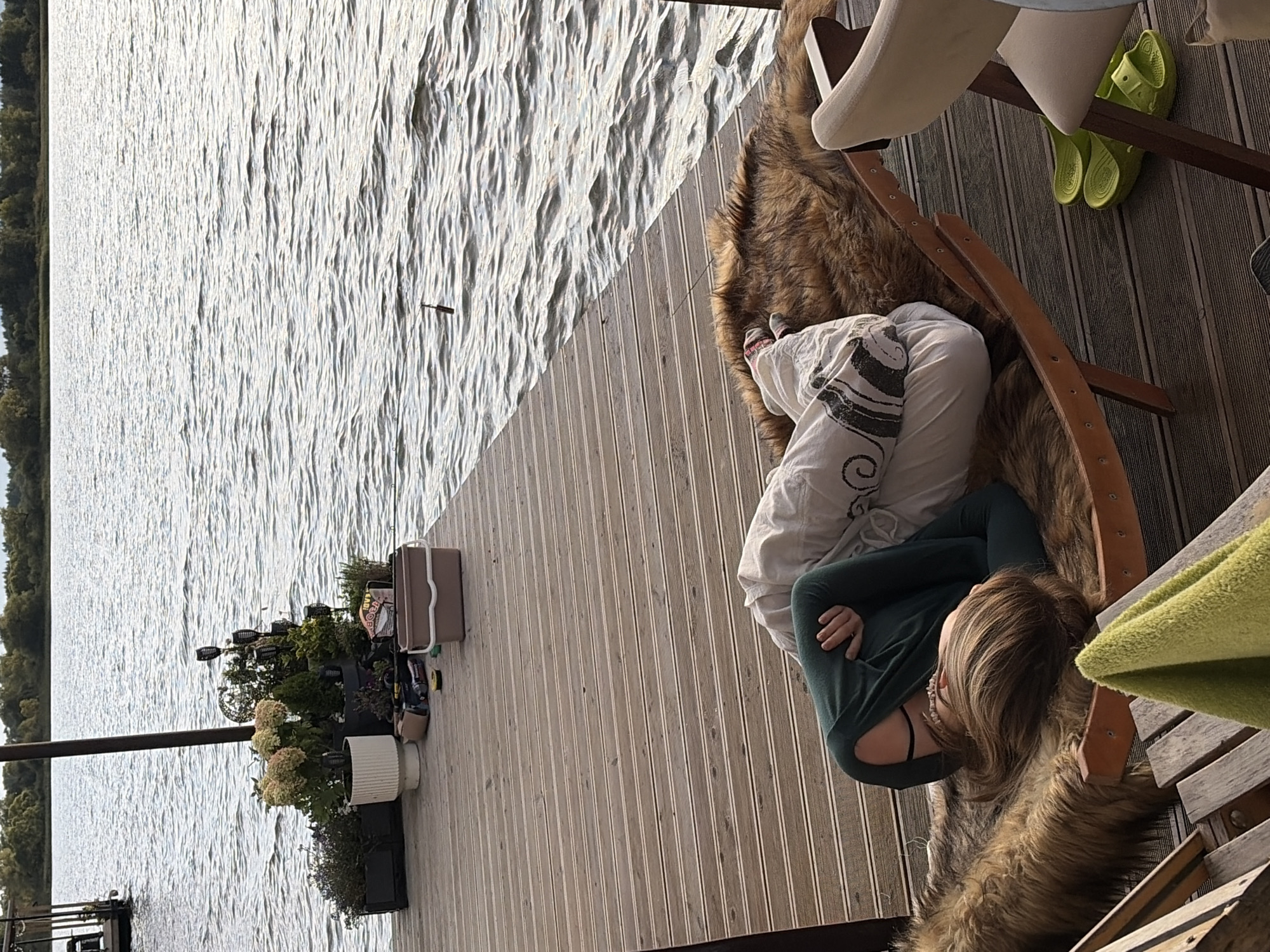 A person lies on a fur-covered lounge chair on a wooden pier in Vidogoshchi, Russia. The individual is wearing a green long-sleeved shirt and white pants, with their head resting on a pillow. The chair is positioned near the edge of the pier, overlooking a body of water. A potted plant and a brown box are visible on the pier near the chair. The water is calm with gentle ripples. The sky is overcast, and trees line the distant shore. A pair of bright green flip-flops are placed on the pier to the right of the chair. A light-colored chair is also present on the right side of the frame.