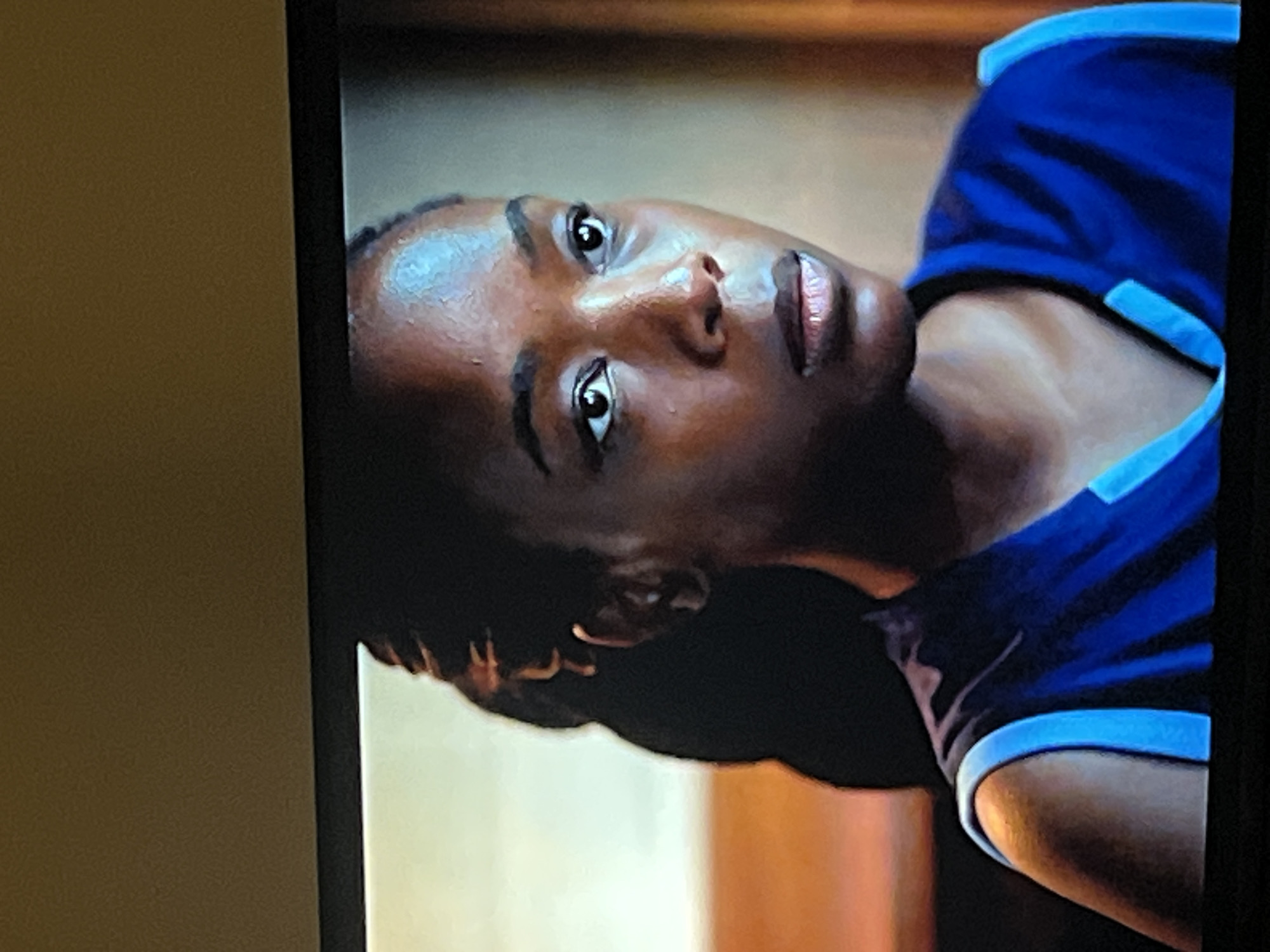 A medium close-up shot shows a Black woman with dark skin and dark hair pulled back. She is wearing a royal blue athletic top with light blue trim. The woman is looking to her right with a neutral expression. Her skin is subtly illuminated, highlighting her cheekbones and forehead. The background is softly blurred, with warm tones of orange and brown, suggesting an indoor setting with warm lighting. There is no indication of a specific location in Gura Top, Nigeria.