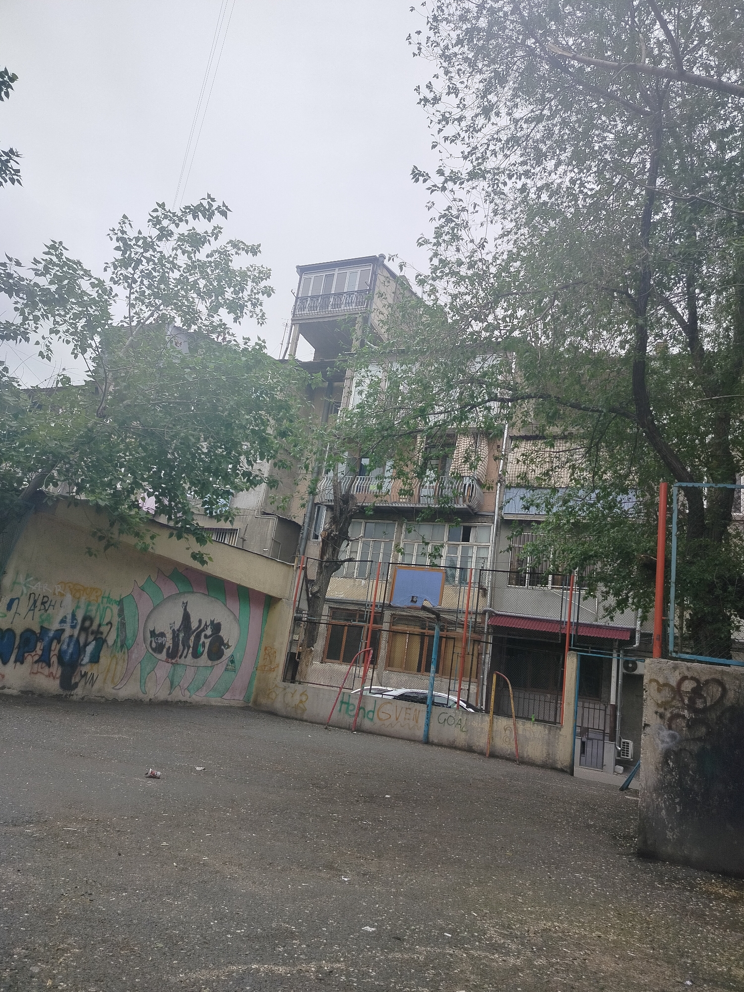 A somewhat rundown urban scene, likely in Yerevan, Armenia, shows an open courtyard with a textured gray asphalt surface. The scene is dominated by a building complex featuring a mix of architectural styles and varying states of repair. Some sections of the building have balconies. The building's exterior shows signs of age and weathering. A perimeter wall features colorful graffiti and murals. A playground element (basketball hoop and goal) is situated next to the building. Trees are present, framing the scene. Overhead power lines traverse the top portion of the image. The scene is overcast. A car is parked behind the fence.