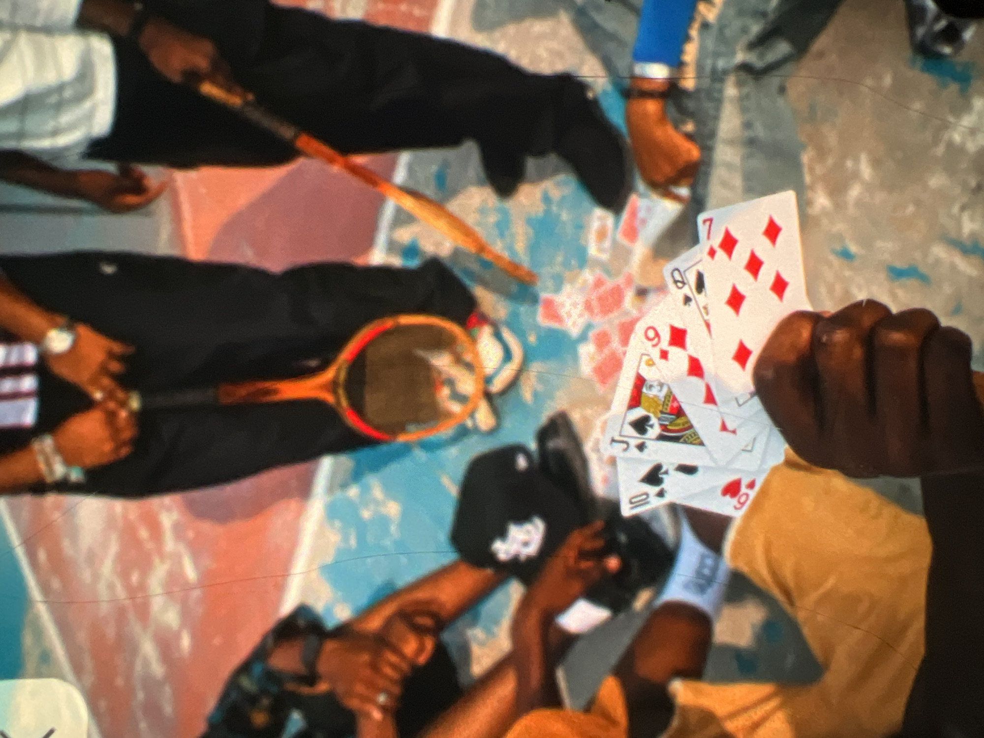The image depicts a group of people, seemingly engaged in a game of cards, on a court. The court surface is visible, with areas of red and blue. In the foreground, a hand is holding a set of playing cards, featuring spades and diamonds. Some of the cards clearly display numbers like "10" and "6," and face cards such as a "J." Scattered on the ground are more playing cards, some face up and others face down.
One person, positioned towards the center of the image, is holding a tennis racket with an orange frame. This individual is wearing dark pants and a white and black striped shirt. Another person, on the right, is dressed in black pants and a white shirt, also holding a tennis racket.
A black baseball cap with a white logo is placed on the ground near a person's knees. The people's skin tones are dark, suggesting they are likely of African descent. The lighting appears to be natural daylight. The scene conveys a casual, informal gathering, possibly in an outdoor recreational area. The specific location in Lagos, Nigeria, cannot be determined from the visual information alone.
