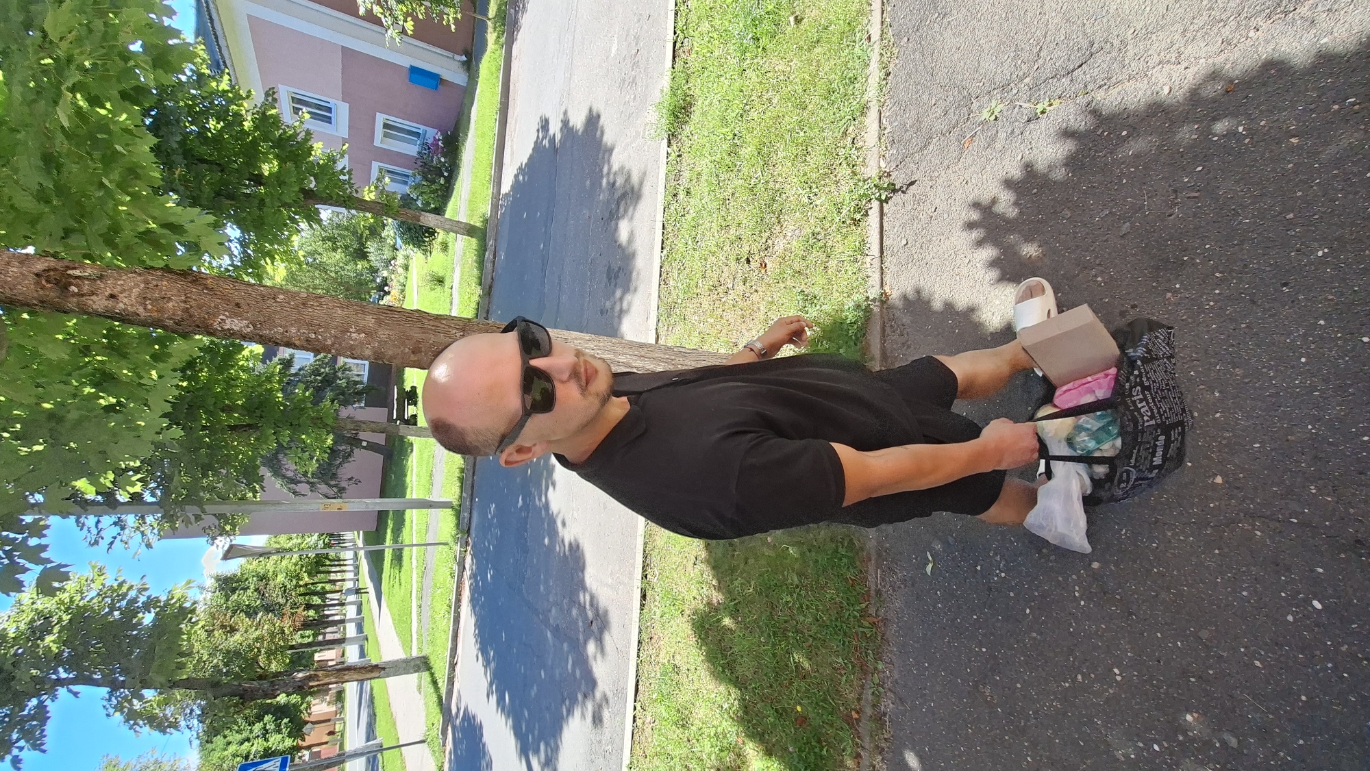 A man with a shaved head and sunglasses walks on a paved sidewalk next to a tree. He is wearing a black polo shirt and black shorts. He is carrying a black tote bag filled with various items. The tote bag has the word "Paris" printed on it in white letters. He is wearing white slides on his feet. In the background, there is a street, grass, trees, and buildings. The sky is blue and there are shadows on the ground from the trees. The scene appears to be in a residential area in the city of Myadzyel, Belarus.