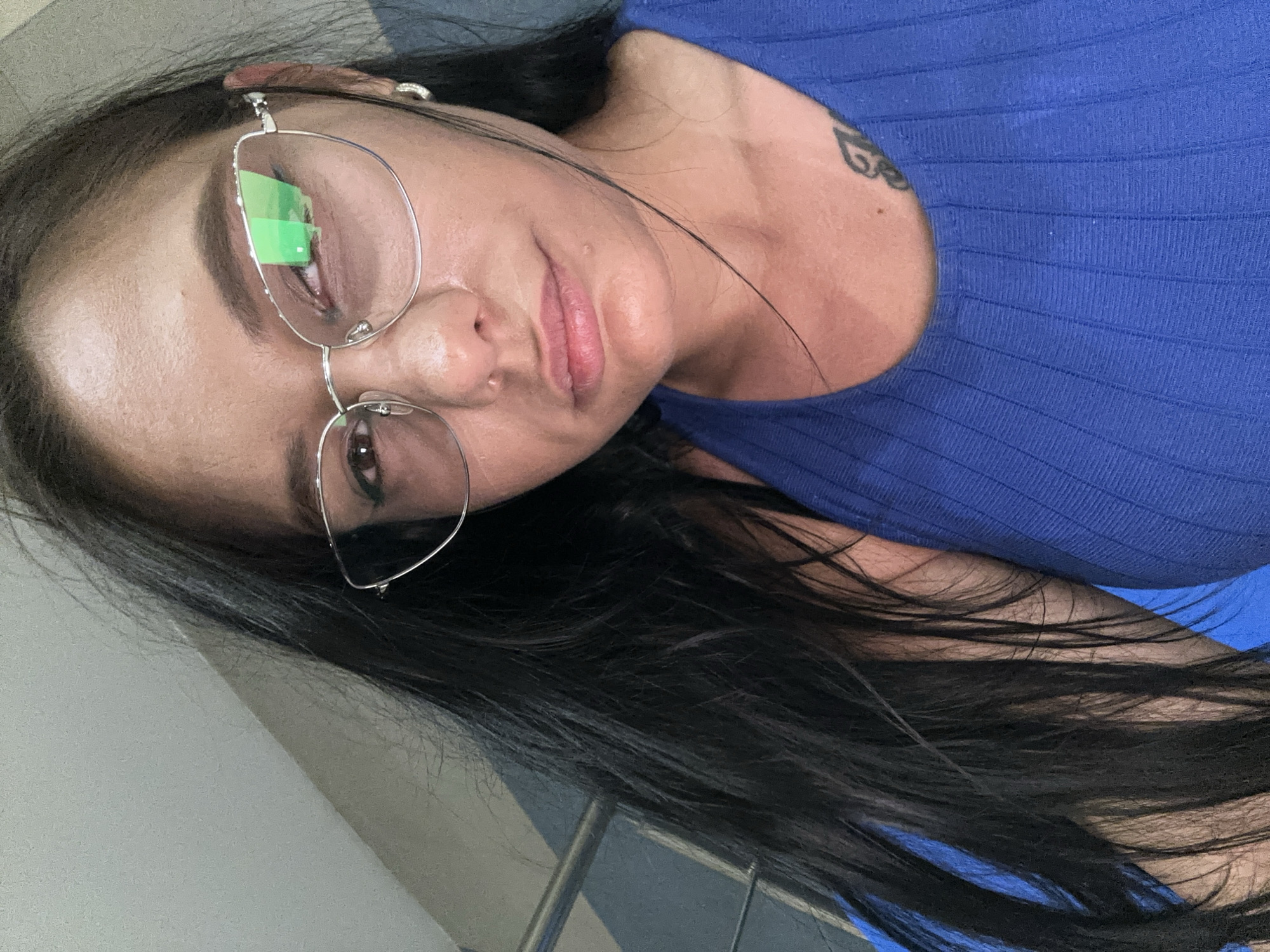 A close-up, high-angle selfie of a woman with long, dark hair and glasses is shown. She is wearing a blue, ribbed tank top. A small tattoo is visible on her chest. The background appears to be an indoor staircase with a light gray wall and a blue railing. The image was taken on a staircase in Sofia, Bulgaria.