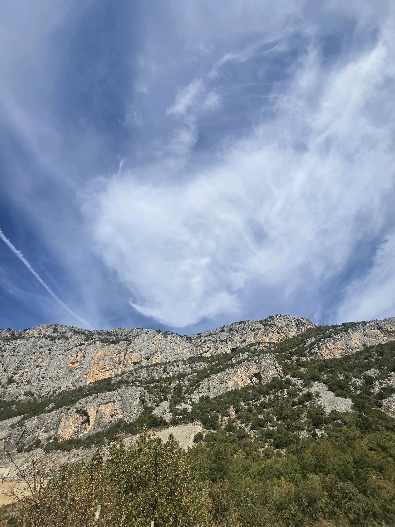 The image shows a mountain range under a bright blue sky with wispy white clouds. The mountains are rocky and steep, with patches of green vegetation clinging to their slopes. In the foreground, there is more dense green foliage. The sky features a prominent contrail from an aircraft. The scene is a natural landscape, likely a mountainous region outside of a city. No people or specific man-made structures are discernible in the image, apart from what appears to be a distant fence line along the mountain slope. The overall impression is one of rugged natural beauty and open sky.
