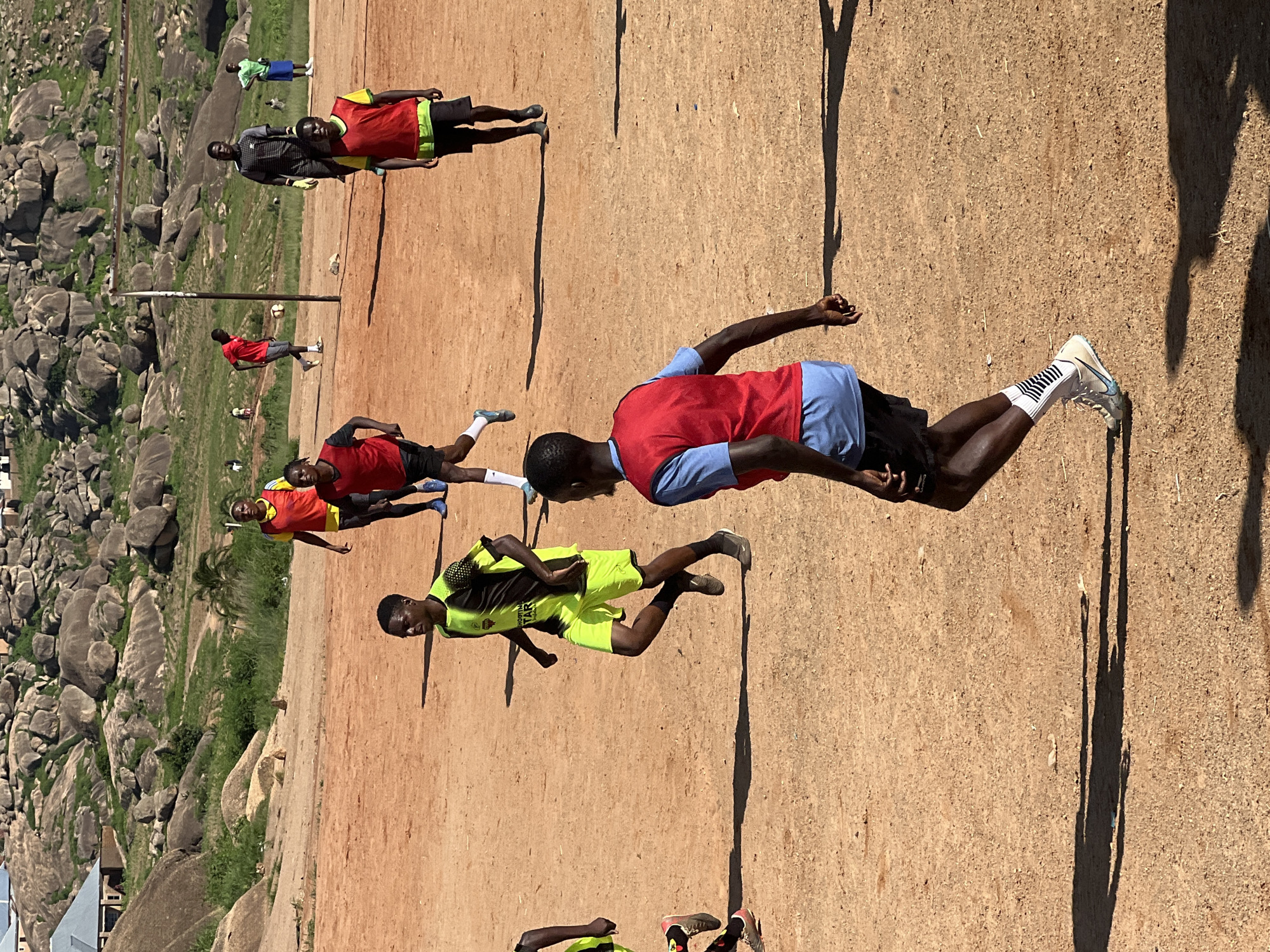 A group of young men, likely a soccer team, are practicing on a dirt field in Jos, Nigeria. The terrain is characterized by a sandy, reddish-brown surface with scattered debris and the shadows of the players and a nearby pole cast on the ground. In the background, a dramatic landscape of large, weathered rock formations rises above green, grassy slopes. Several residential structures, appearing to be simple houses with corrugated metal roofs, are visible nestled among the rocks and vegetation. The players are dressed in various athletic attire, including jerseys, shorts, and cleats, with some wearing bibs over their shirts. Their actions suggest a training session or a casual game, with some individuals running and others appearing to observe or participate. The overall atmosphere is one of informal sporting activity set against a rugged, natural backdrop.