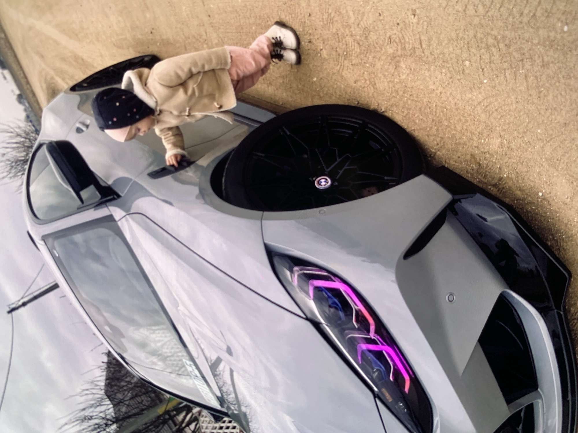 An outdoor, low-angle, eye-level medium shot shows a young child and a car. The car, a grey sedan, is angled towards the viewer's left, with its front facing to the left. The car has custom purple and pink lighting in its headlights. A young child, wearing a black hat with colorful polka dots, a beige coat, pink pants, and white boots, is standing on the right side of the car, with their right hand reaching for the car door handle. The child is leaning into the car slightly, appearing to interact with it. The ground is covered in a dirt or gravel path. In the background, there are bare tree branches and a utility pole with wires. The sky is overcast.