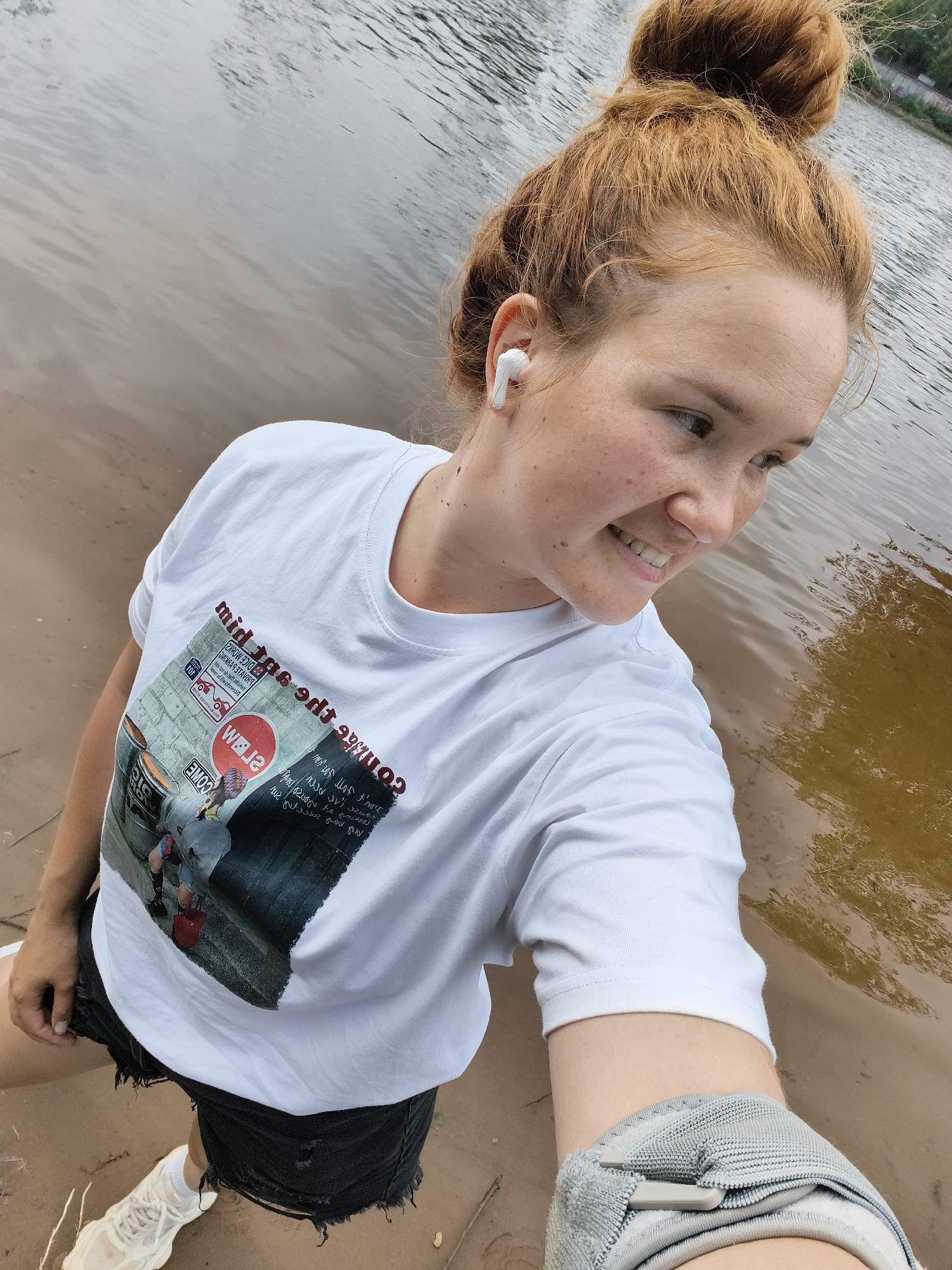 A young woman with red hair pulled up in a bun is taking a selfie by the water. She is wearing a white t-shirt with a graphic design and text, black distressed denim shorts, and white sneakers. She has white wireless earbuds in her ears and is smiling. The background shows a body of water with ripples and a sandy shore. The lighting suggests it is daytime, possibly overcast. Visible text on her shirt includes "mid tas edt 990?", "comes ar PV", and "DON' F JOT JOM KOU". The overall scene suggests a casual outdoor activity.