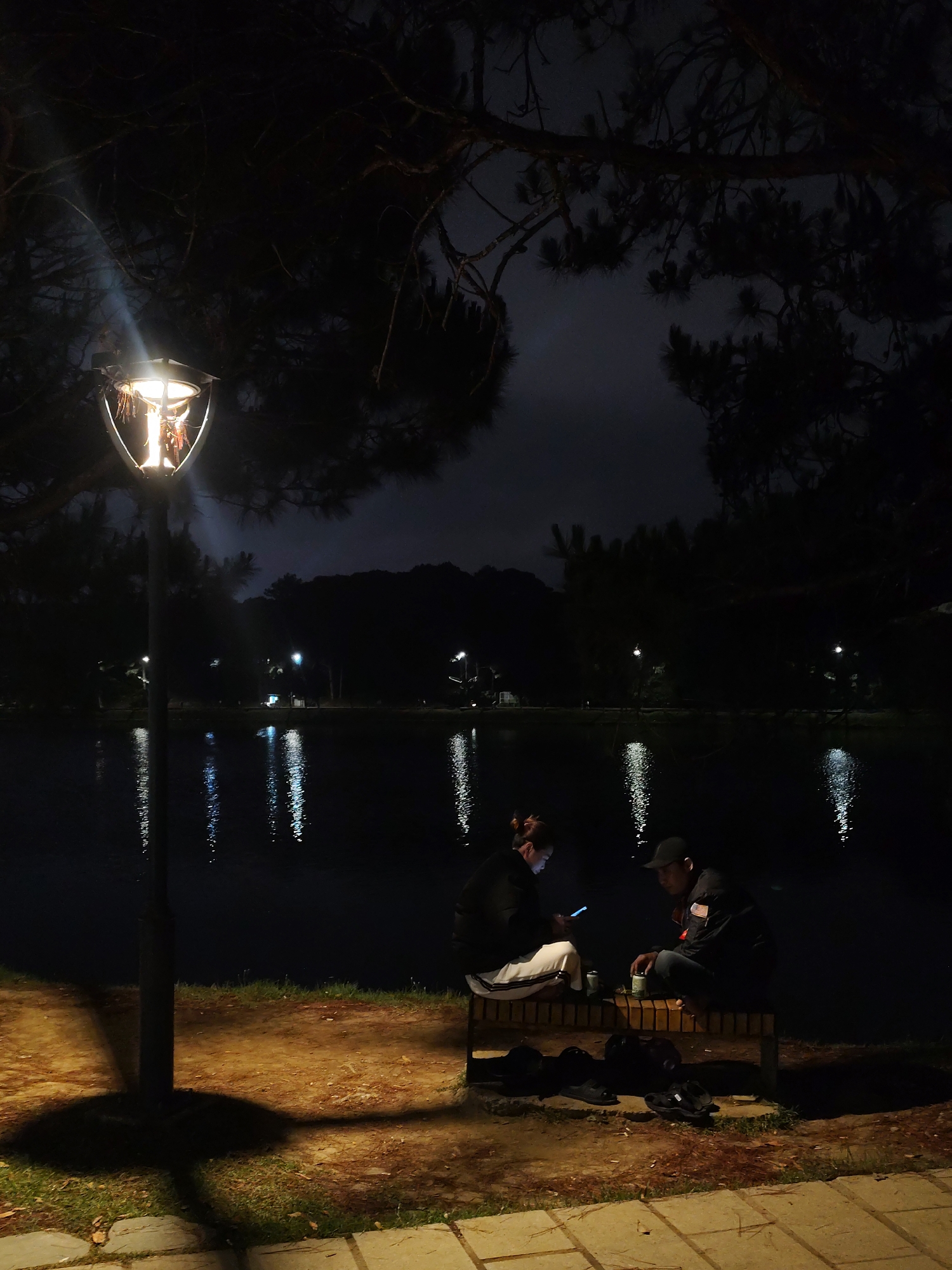 Captured at night, the scene unfolds along the edge of Xuan Huong Lake in Da Lat, Vietnam. A light post illuminated by a glowing bulb casts a warm glow on the area, and is partially obscured by tree branches. Two individuals are seated on a bench near the water's edge, facing each other in low light. One person is looking at their phone. Various pairs of shoes and black bags are placed in front of the bench. Light reflections are visible on the surface of the lake, with trees and hills forming a dark backdrop in the distance.
