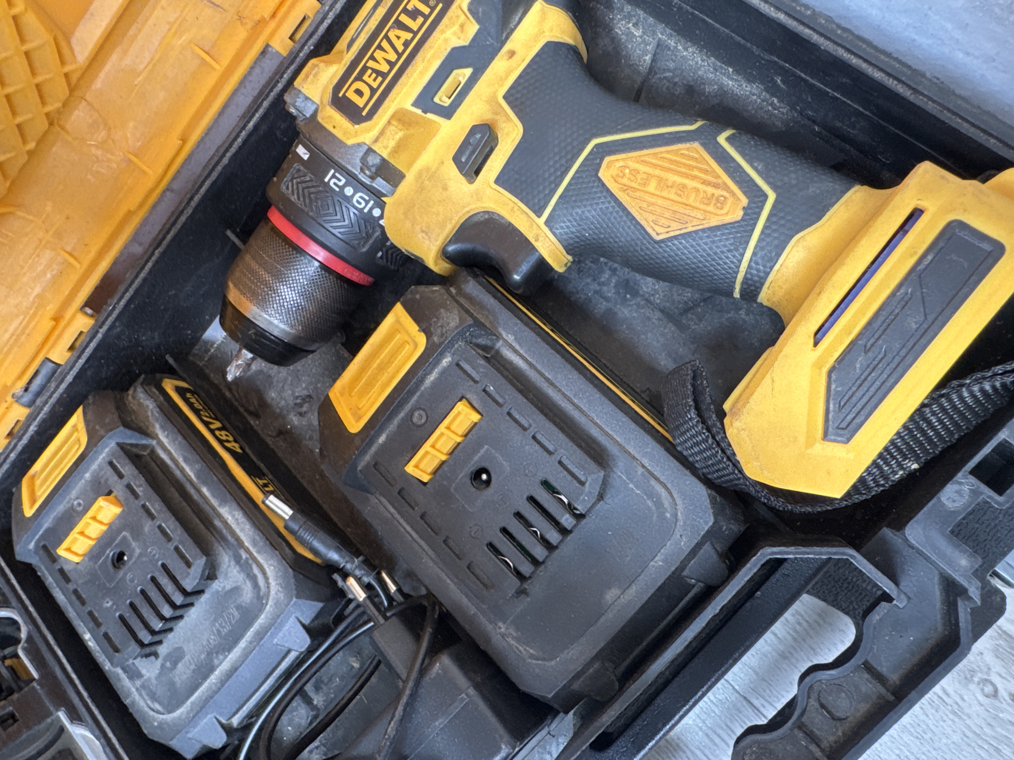 The image shows a black and yellow DeWalt cordless drill and two batteries inside a black and yellow plastic carrying case. The drill has a red ring around the chuck, and the word "DEWALT" is visible on the side. One of the batteries is labeled "48V 2.0AH". The carrying case is open, and the interior is visible. The background is out of focus, but appears to be a wooden floor. The scene is likely a workshop or garage. There is no information available about the location in the city of Яромінскі сельскі Савет, Belarus.