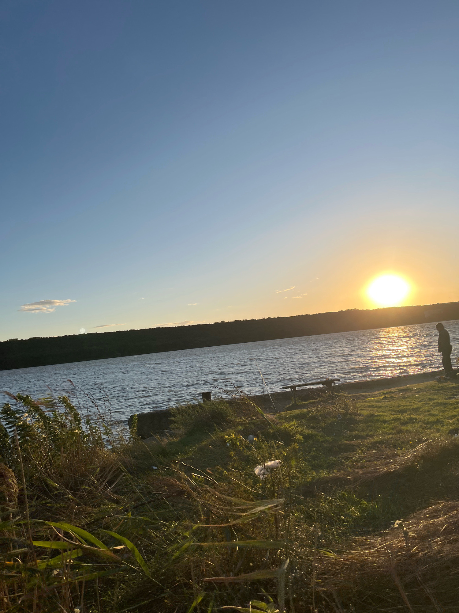 The image depicts a tranquil sunset scene by a body of water, likely a lake or river, with a treelined horizon. In the foreground, tall grasses and vegetation grow along the grassy bank. A single person stands near the water's edge, facing away from the camera and towards the setting sun. The sun itself is a bright orb, casting a shimmering reflection on the water's surface. The sky is clear blue, with a few wispy clouds. The overall atmosphere is peaceful and serene. The location is identified as Korzhevo, Moldova.