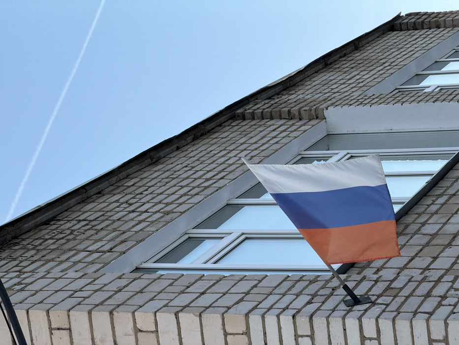 A section of a light-colored brick building is depicted against a clear blue sky in the city of Chaikovsky, Russia. A Russian flag, exhibiting vertical stripes of reddish-orange (left), blue (middle), and white (right), is mounted horizontally from a black L-shaped metal bracket. The bracket is affixed to the light-toned brick wall, positioned between two white-framed windows. The flag extends outward from the building, appearing slightly creased. The brickwork features uniform, light-colored bricks with darker mortar lines. A dark, narrow overhang or roofline runs along the top edge of the building. The sky is a uniform bright blue, with a single thin white contrail visible diagonally across the upper right quadrant. No individuals or specific actions are present within the frame.