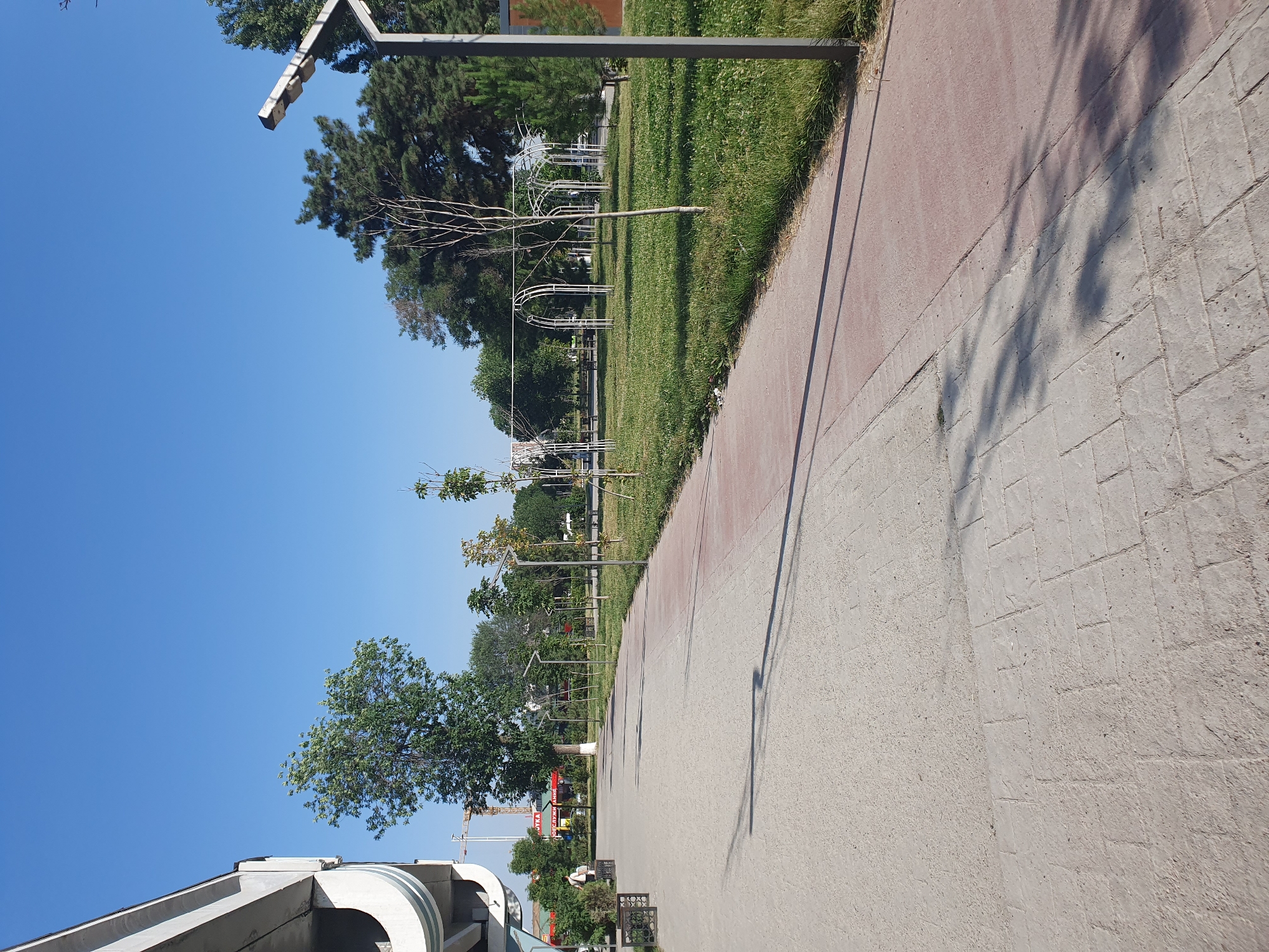 The image depicts an outdoor scene on a clear day, likely in Tashkent, Uzbekistan. In the foreground, a wide paved walkway with a reddish border curves to the right. The walkway is composed of lighter-colored paving stones and is partially in shadow. To the right of the walkway is a grassy area with scattered trees and decorative metal arches. Several modern streetlights are visible along the path and in the grassy area. In the background, there are more trees, some buildings, and a construction crane. On the left side of the walkway, a concrete structure, possibly part of an overpass or building, is visible. A single person is walking on the walkway in the distance. The overall impression is of a park or public space in an urban environment.