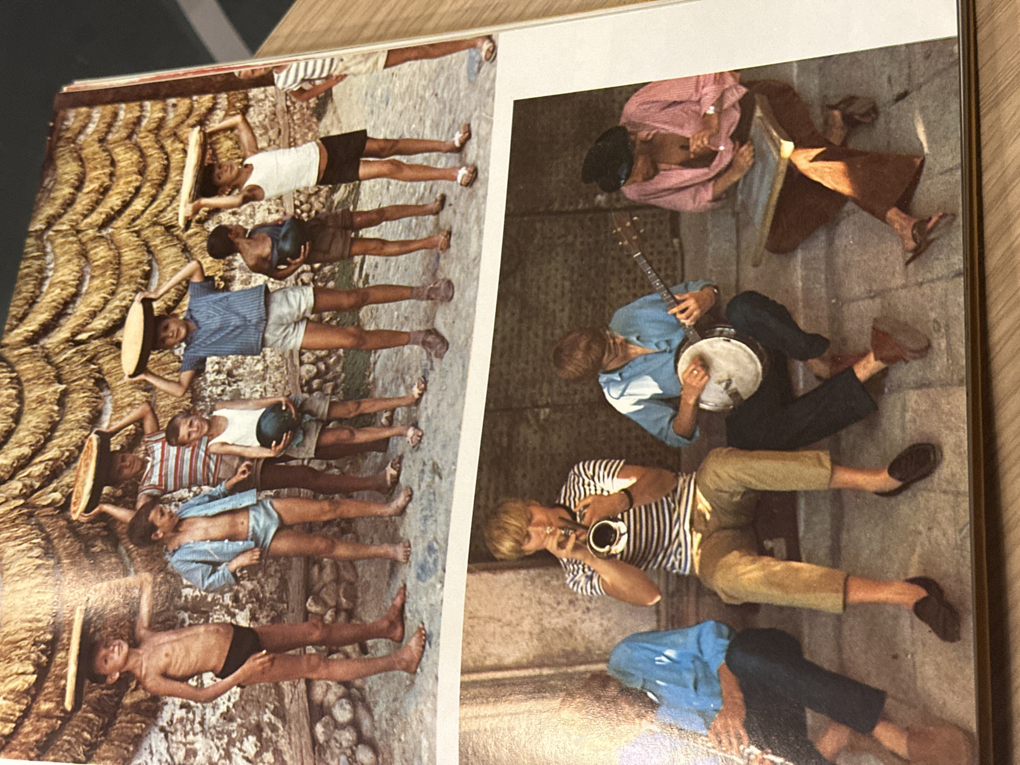 The image depicts two adjacent pages of a magazine or book. On the left page is a photo of a group of young people lying down on what appears to be rough terrain, possibly a dried river bed or a beach with coarse sand and pebbles. They are wearing a variety of clothing, some are bare-chested, while others are wearing shirts and shorts. Several are holding objects above them, such as a hat or a ball.
The right page features a photograph of a group of musicians lying down on a paved surface, presumably a street or square. They are playing musical instruments, including a banjo, a trumpet, and possibly another instrument that is obscured. The environment looks like a paved street or sidewalk.
The location cannot be determined to be in Novi Sad, Serbia, with the given visual information.