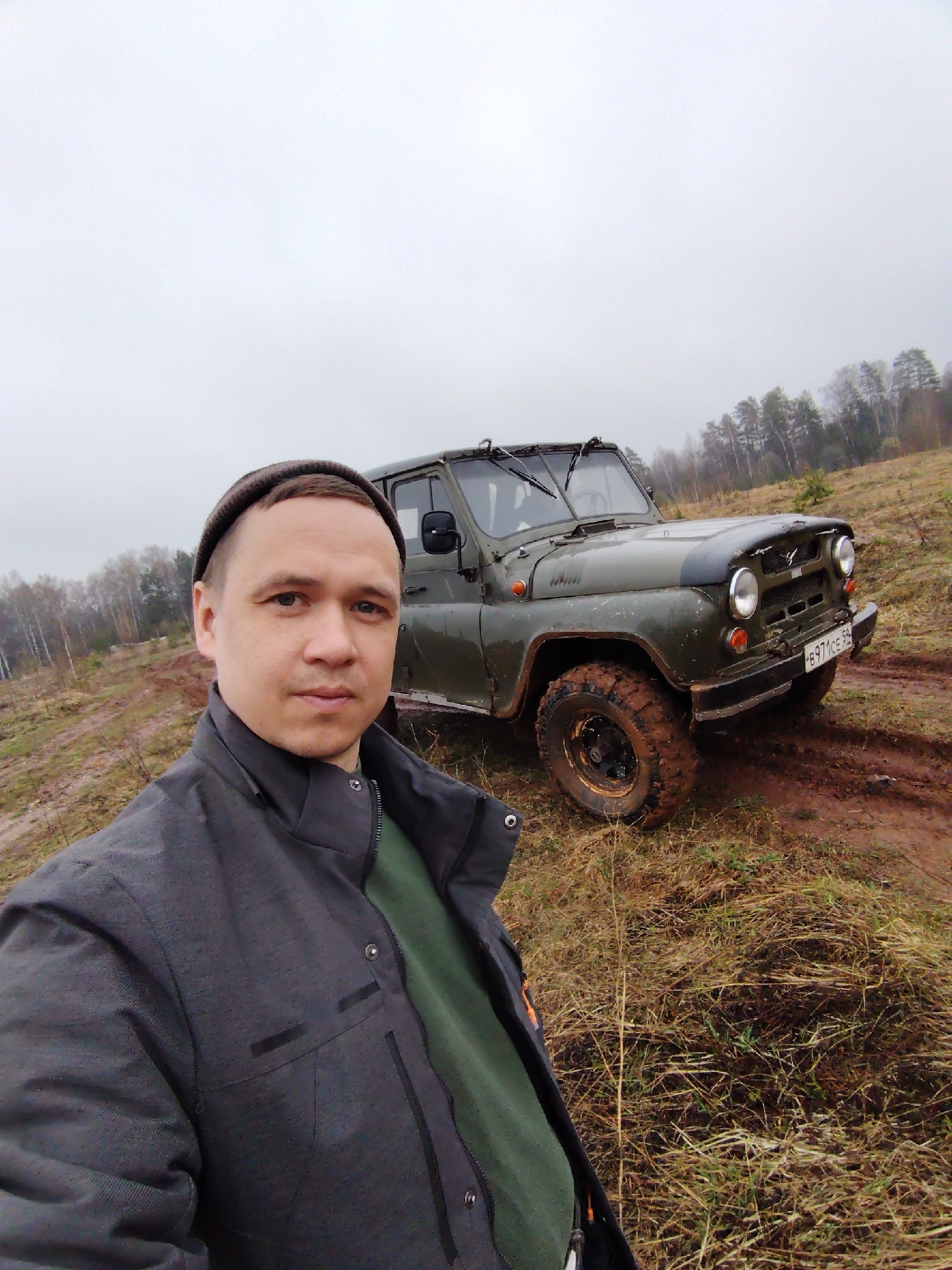The image depicts a man standing outdoors next to an off-road vehicle, possibly a UAZ-469, in a muddy field in Марковский, Russia. The man is wearing a dark grey jacket, and a beanie, and the vehicle is dark green with mud-splattered tires. The terrain appears to be wet and overgrown with dry vegetation, and there's a line of bare trees in the background under a grey sky. The vehicle has the license plate number B971CE 59.