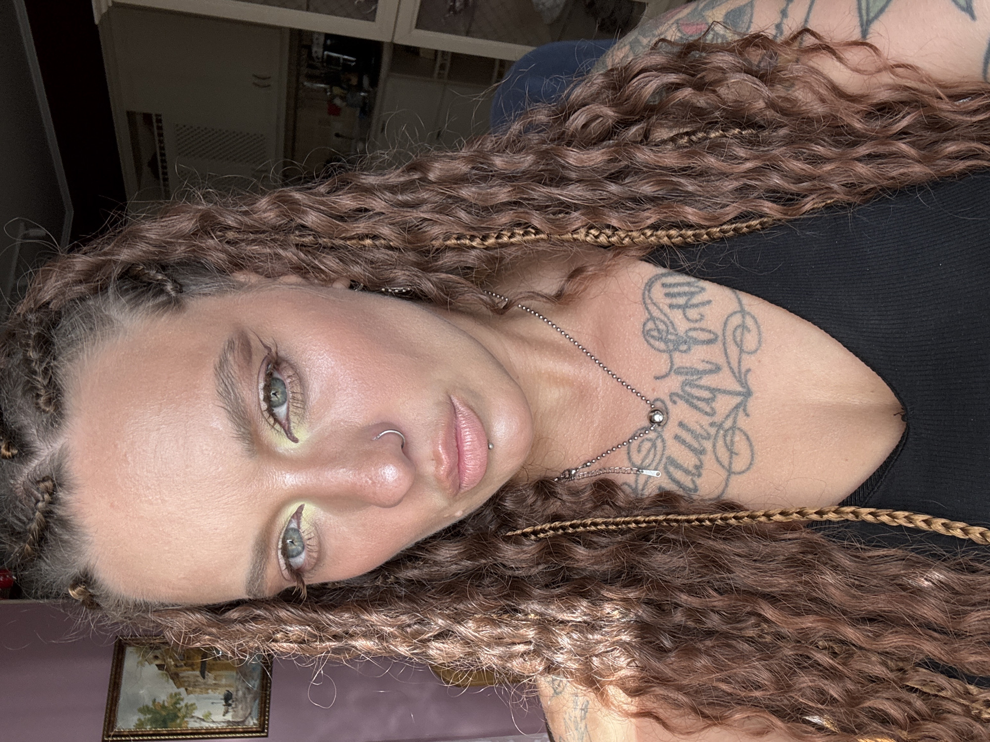 This is a portrait-style, medium shot of a young woman lying on her side. She has long, curly, dark brown hair with lighter brown braided accents. Her face is turned towards the camera, and she has striking blue eyes with pale green eyeshadow. She is wearing a silver necklace with a circular pendant and a small zipper charm. She has a nose ring and a lip piercing. There is a tattoo visible on her chest and upper arm, with stylized lettering. The background appears to be an indoor setting with cabinets or shelves visible in the upper portion of the frame, and a framed picture on the wall in the lower left. The lighting suggests it might be daytime, with some artificial light source contributing to the overall illumination. The mood is somewhat relaxed or contemplative. There is no visible text in the image.