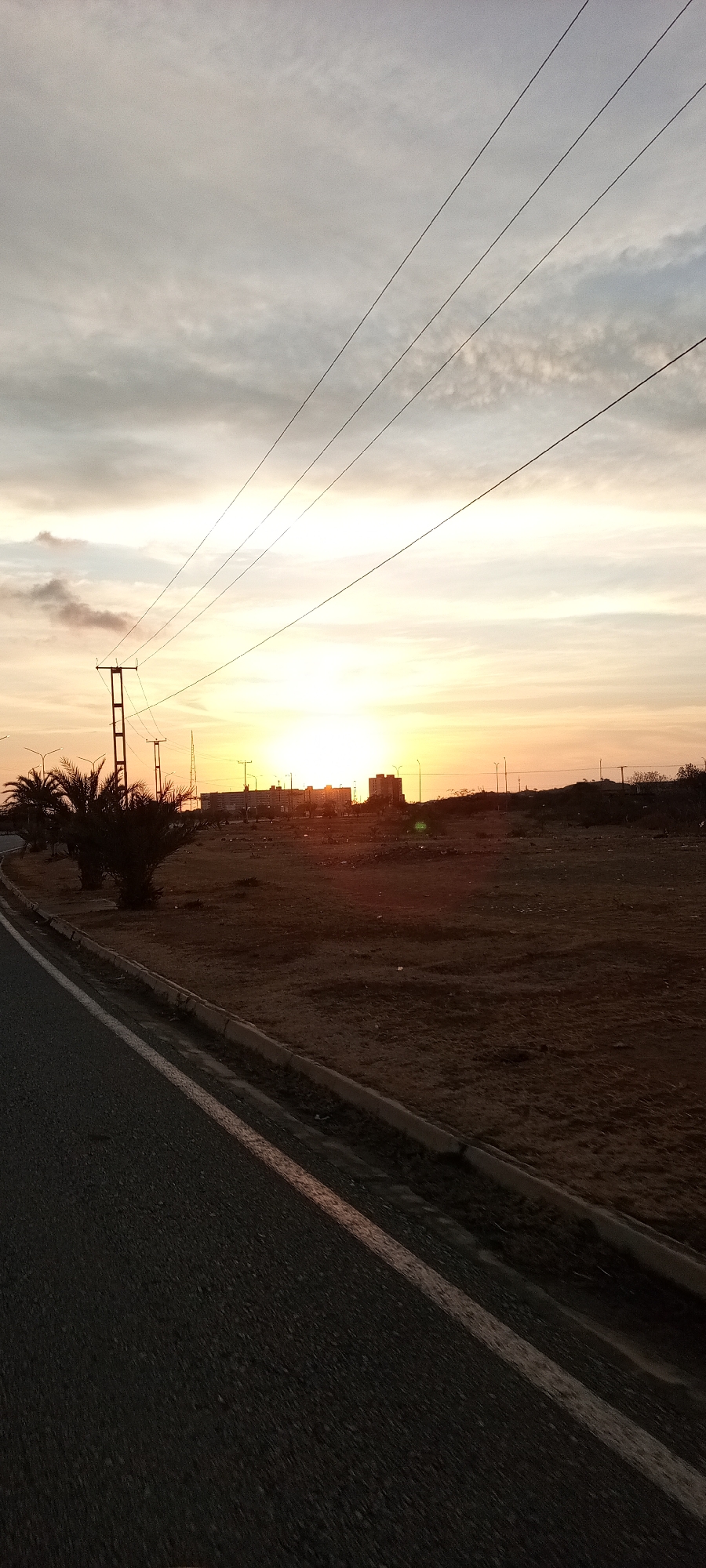 The image shows a sunset scene on the outskirts of a city. The sun is setting in the distance, casting a warm glow over the landscape. In the foreground, there is a paved road with a curb. Beyond the road is a barren, dirt-covered area with scattered debris. Several palm trees are visible along the roadside. In the distance, there are buildings and lampposts silhouetted against the setting sun. Power lines stretch across the sky. The overall impression is one of a desolate, undeveloped area on the edge of an urban environment.