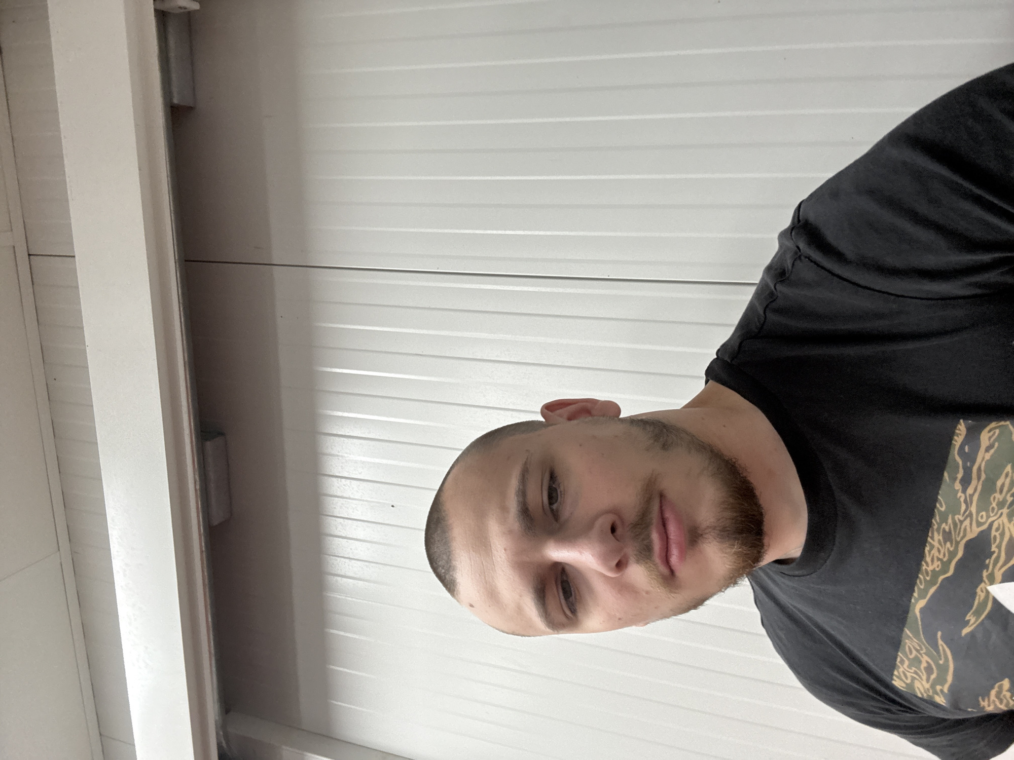 A young adult man is depicted in a close-up, chest-up portrait, with the image rotated 90 degrees clockwise. He has a buzz cut hairstyle and a short, neatly trimmed beard, looking directly at the camera with a neutral expression. He is wearing a black t-shirt that features a distinctive green, brown, and black camouflage pattern, possibly a tiger stripe design, visible on his right shoulder and chest.
The background is dominated by a white, horizontally ridged surface, suggestive of industrial paneling or a roll-up garage door. On the far left, a vertical white beam or structural element is present. In the upper left background, there are hints of metallic mechanisms or tracks, consistent with a garage or a similar utility space. The lighting is even, likely artificial, illuminating the scene. The setting for this image is Stara Zagora, Bulgaria. No discernible text is visible on his clothing or in the background.
