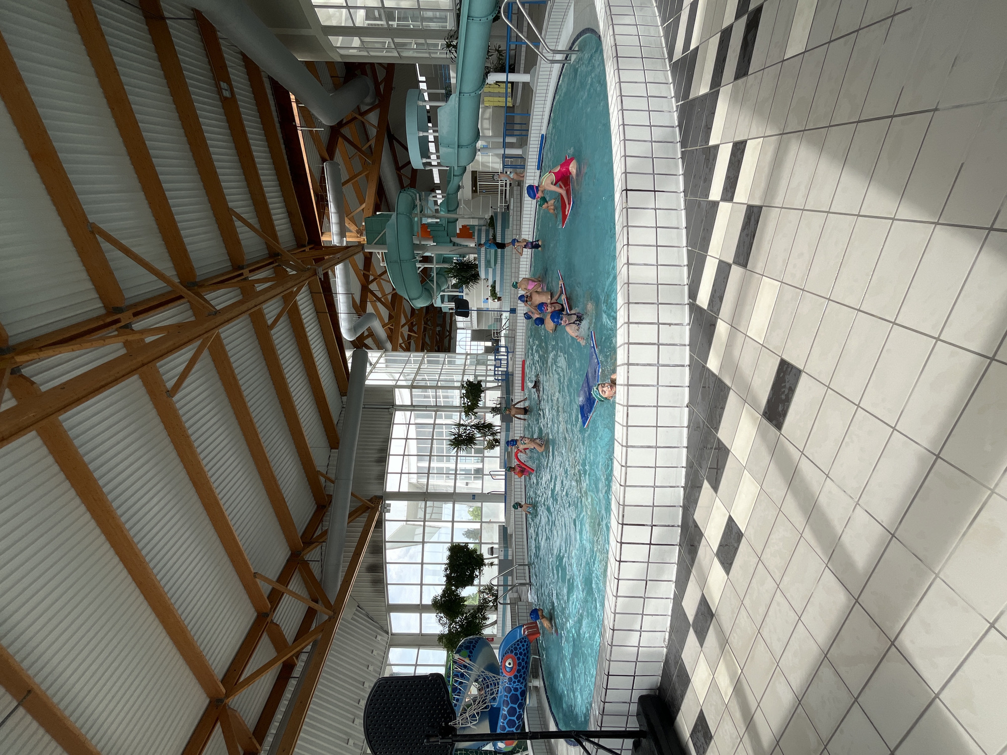 This indoor scene depicts a swimming pool area. The pool is filled with several people of varying ages, presumably swimming or playing in the water. Some are wearing swim caps and using floats or kickboards. A water slide structure is visible at the back of the pool area, suggesting recreational facilities. The pool is surrounded by white tiled walls, with a black and white checkered pattern on the floor. Large windows provide natural light, with indoor plants situated along the window line. Above, the ceiling features a wooden beam structure supporting the roof. A basketball hoop stands near the pool. This scene likely takes place at a recreational facility in Andrézieux-Bouthéon, France.
