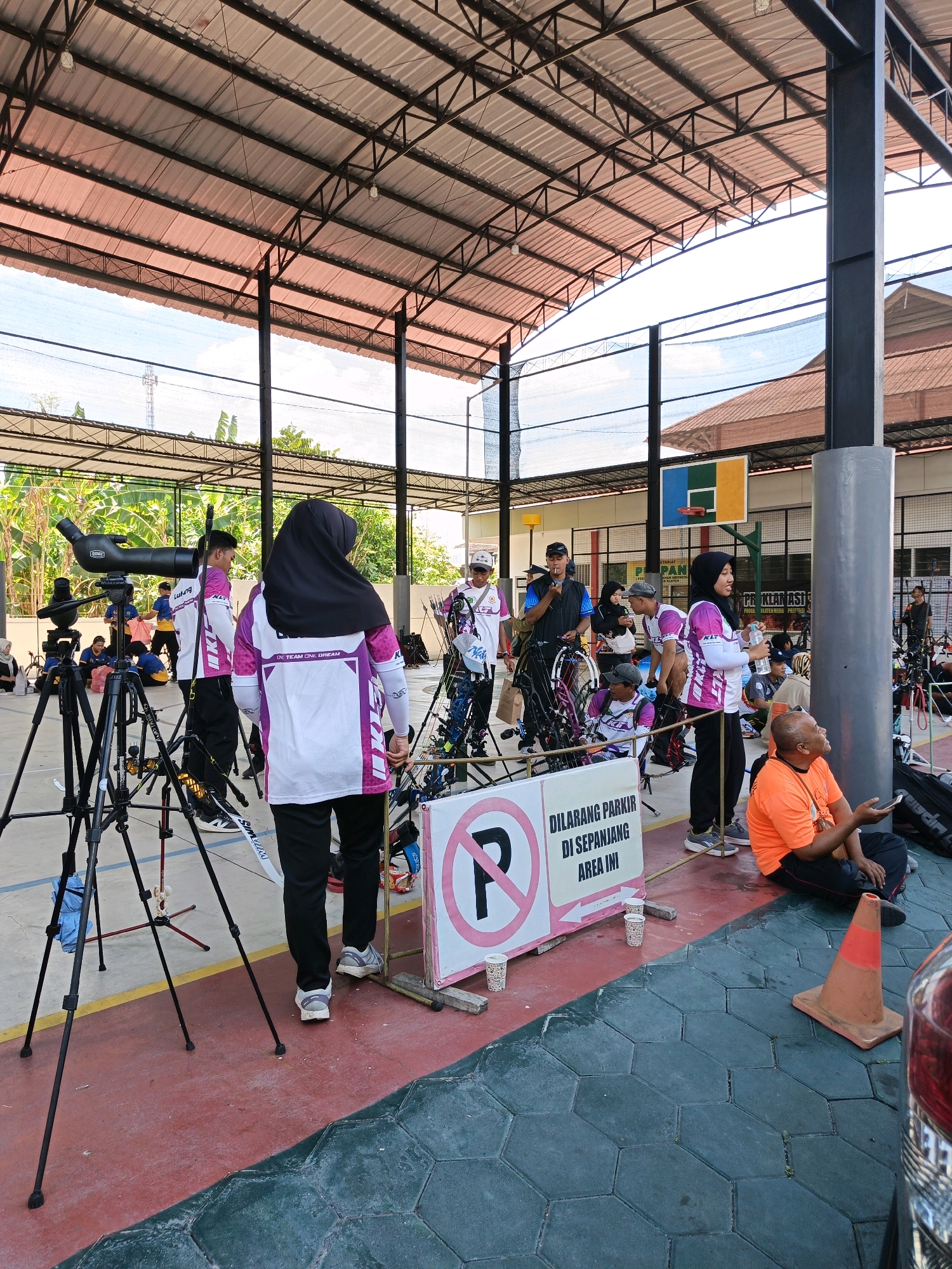 The image captures an outdoor archery event at what appears to be a sports complex in Jonggrangan, Indonesia. A covered structure with a metal roof supported by dark gray pillars provides shade over a red and green tiled floor. In the foreground, a woman wearing a hijab and a white and purple tracksuit stands facing away from the camera, holding a water bottle. To her left, a tripod-mounted telescope is positioned, and beyond it, several individuals are seen preparing for or observing the archery competition. Archery equipment, including bows and arrows, is visible. A prominent sign with a pink circle and a diagonal line over a letter 'P' indicates "DILARANG PARKIR DI SEPANJANG AREA INI," meaning "NO PARKING ALONG THIS AREA." In the background, a basketball hoop is mounted on a wall. On the right side of the frame, a man in an orange shirt is seated on the ground, looking at his phone. The overall atmosphere suggests an organized sporting event with participants and spectators gathered under the covered area.