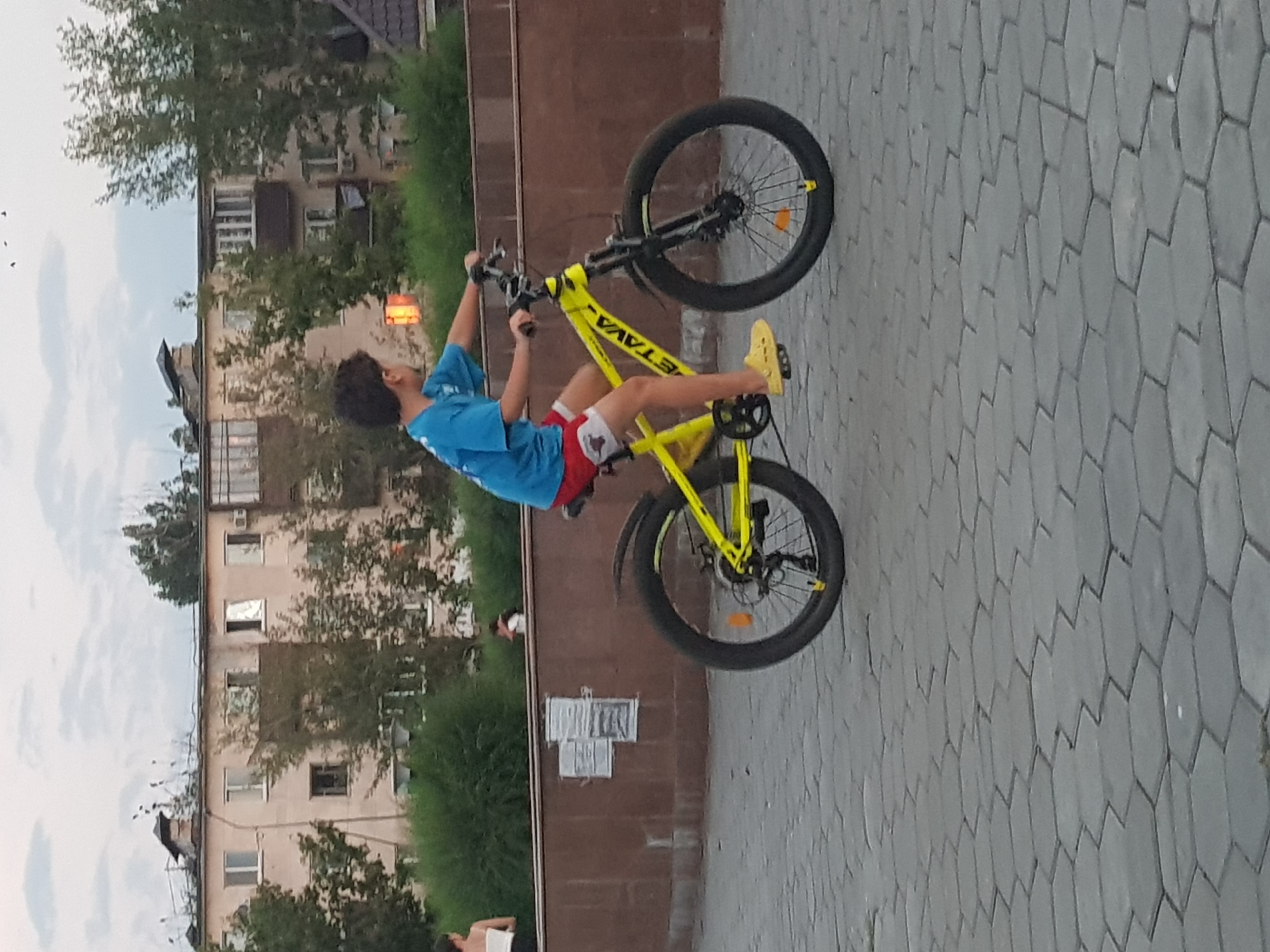 A young boy rides a bright yellow bicycle with thick tires on a hexagonal paved pathway in the city of Kurchatov, Kazakhstan. The boy is wearing a blue t-shirt and red shorts. He has dark curly hair and is looking forward as he pedals. In the background, there is a multi-story building with several windows and balconies. Trees and greenery are visible in front of and around the building. A brown wall runs horizontally across the mid-ground, behind which is a dense hedge of green bushes. The sky above is overcast.