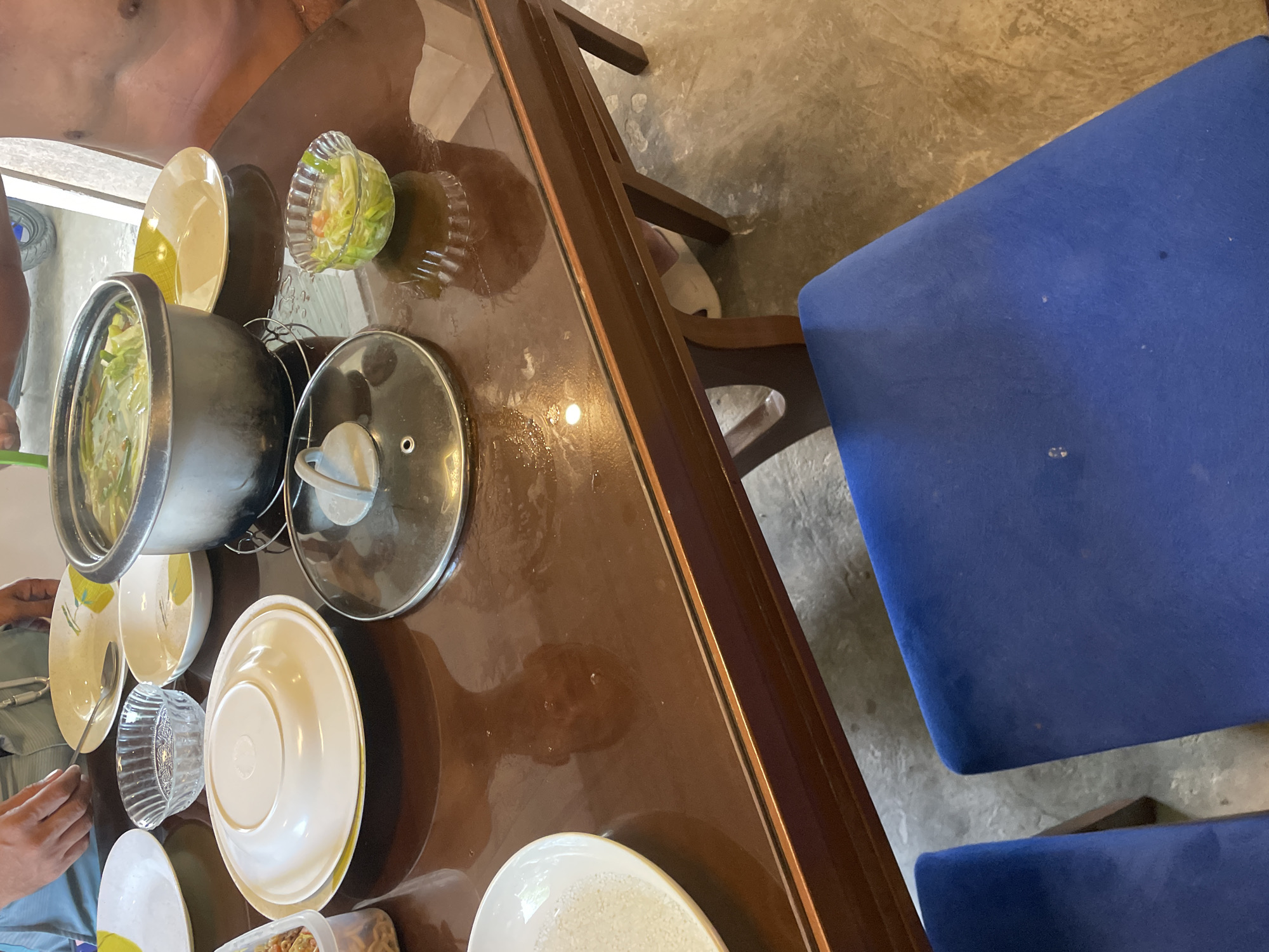 The image depicts a dining table laden with dishes, a pot, and serving bowls. A person is partially visible on the left side of the frame, along with their hand. A person with visible skin and clothing is at the top left of the frame. The table has a glossy brown surface and is surrounded by at least two blue upholstered chairs. The location is Hijo, Philippines.
