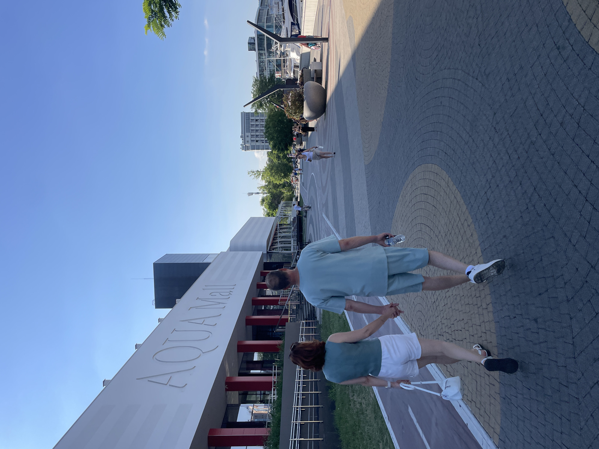 A couple is walking hand-in-hand on a paved walkway in front of a modern building complex that reads "AQUA Mall" in large letters. The woman, on the left, is wearing a green sleeveless top and white shorts. The man, on the right, is wearing a light blue t-shirt and matching shorts. He is holding a bottle of water in his right hand. The walkway is made of patterned pavers, with a red and white striped bike path running alongside it to the left. In the background, beyond the Aqua Mall, there are more buildings, trees, and a body of water with boats docked. The sky is clear and blue. The scene appears to be in a waterfront area of a city. The location is Maiorovskii, Russia.