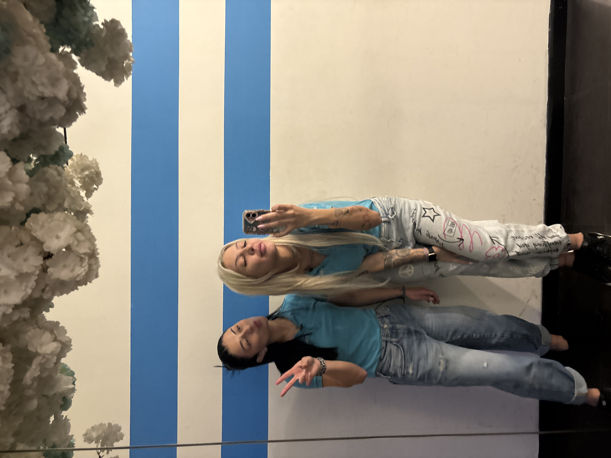 Two women pose for a selfie in front of a white wall with vertical blue stripes, located next to a floral display and a black door. The first woman, with long blonde hair, holds a phone and wears a blue t-shirt and light-colored sweatpants with writing. The second woman, with long dark hair, makes a peace sign and wears a blue t-shirt and jeans. The photo was taken indoors, likely in Plovdiv, Bulgaria.