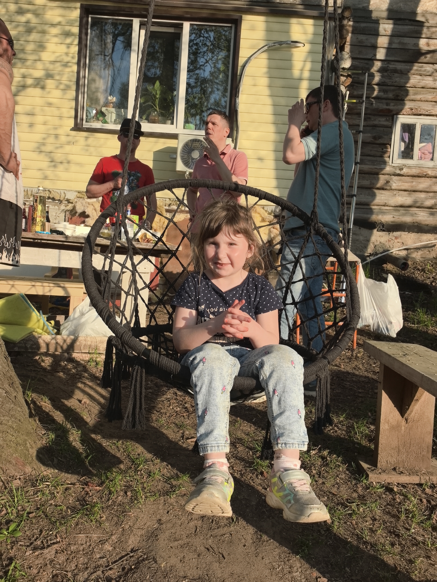 The image shows a young girl sitting in a black hammock-like swing outdoors in a yard. The girl is wearing a dark blue shirt with white polka dots, light blue jeans with ice cream cone patterns, pink socks, and colorful sneakers. She is smiling at the camera with her hands clasped in front of her.
Behind her, several adults are standing near a yellow-sided building with a window. There's a table with various items on it, including bottles and food. The area appears to be grassy with trees and a wooden bench nearby.
The setting suggests a casual outdoor gathering, possibly a family event or barbecue. The location is Noviy, Russia. The presence of people indicates a social interaction.