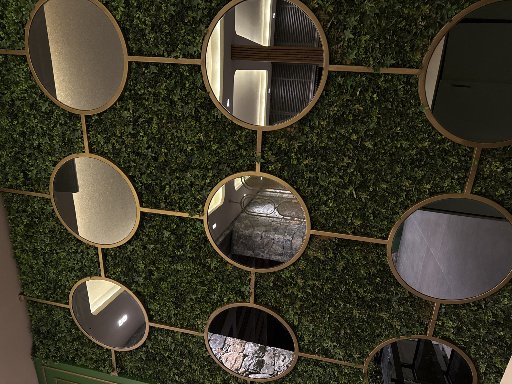 A wall decorated with artificial greenery features a geometric arrangement of circular mirrors with gold frames. The mirrors reflect different parts of an indoor environment. One mirror reflects a ceiling with recessed lighting and what appears to be a wooden slatted partition. Another reflects a stone wall. A third reflects a floor with visible tile lines. Two other mirrors reflect dark, indistinct areas, possibly furniture or cabinets. The scene is well-lit, suggesting it is indoors. There are no people visible in the image. The style of the decor, particularly the artificial greenery and gold accents, suggests a modern and possibly upscale establishment. It is located in Bodrum, Turkey, suggesting a context of a hotel, restaurant, or retail space in a popular tourist destination known for its aesthetic appeal.
