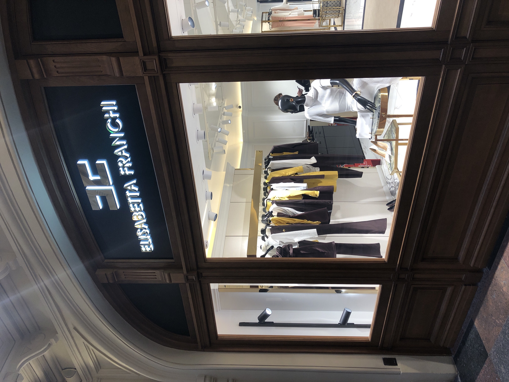 The image shows the exterior of an Elisabetta Franchi store, likely in a European city given the architectural style and the brand's origin. The storefront features a dark wood frame around a display window. The store name, "ELISABETTA FRANCHI," is illuminated in a vertical arrangement, positioned next to the brand's logo. Inside the window, clothing is displayed on hangers, featuring a palette of white, brown, and gold. A black mannequin wearing a white striped sweater is also visible. Overhead, numerous track lights illuminate the interior. The surrounding architecture suggests an upscale retail location. The information about Moscow, Russia cannot be confirmed from the image.
