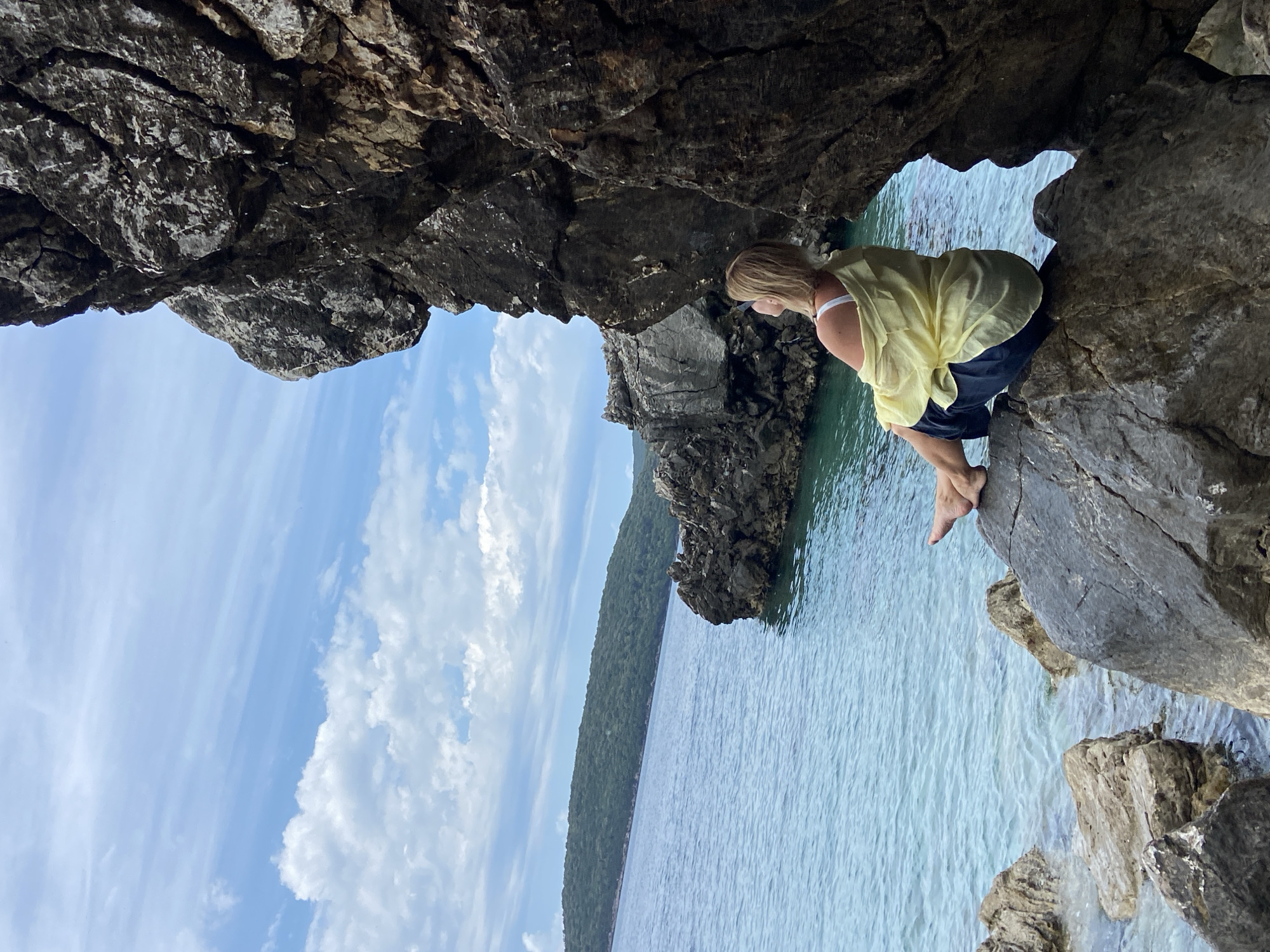 A woman is perched on a rocky outcrop overlooking the Adriatic Sea in Prijevor, Montenegro. The woman is seen from the side, sitting on the rocks, wearing a light yellow shirt and dark shorts. She is looking out at the water. Above and around her are rock formations, forming a natural frame. In the background, the water stretches out to meet a landmass covered in dense green trees. The sky is blue with scattered white clouds. The scene is well-lit, suggesting a bright, sunny day.