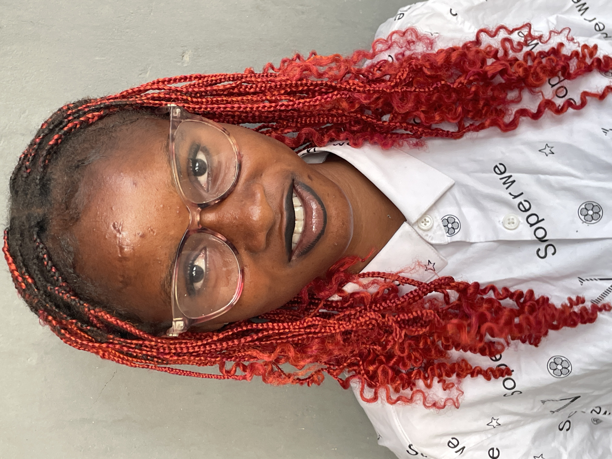 A portrait-style image, rotated 90 degrees counter-clockwise, features a person with dark skin against a plain, light grey background. The subject is likely a woman, looking directly at the camera with a subtle smirk or smile. She wears clear-framed glasses that partially obscure her dark eyes.
Her hair is styled in distinctive braids, a combination of dark and vibrant red strands. Many of the red braids have curly or kinky ends, creating a textured and voluminous effect, suggesting a "boho" or "goddess" braid style.
The person is wearing a white collared shirt with several small, repeating black patterns. These patterns include five-pointed stars and a circular logo containing an abstract design. The word "Soperwe" is also visibly printed on the shirt in black, repeated several times, oriented to be legible when the shirt is worn normally.
The lighting is even, highlighting the subject's features and the details of her hair and clothing. The overall impression is a casual but striking portrait. No specific time of day or outdoor weather conditions can be discerned from the image.