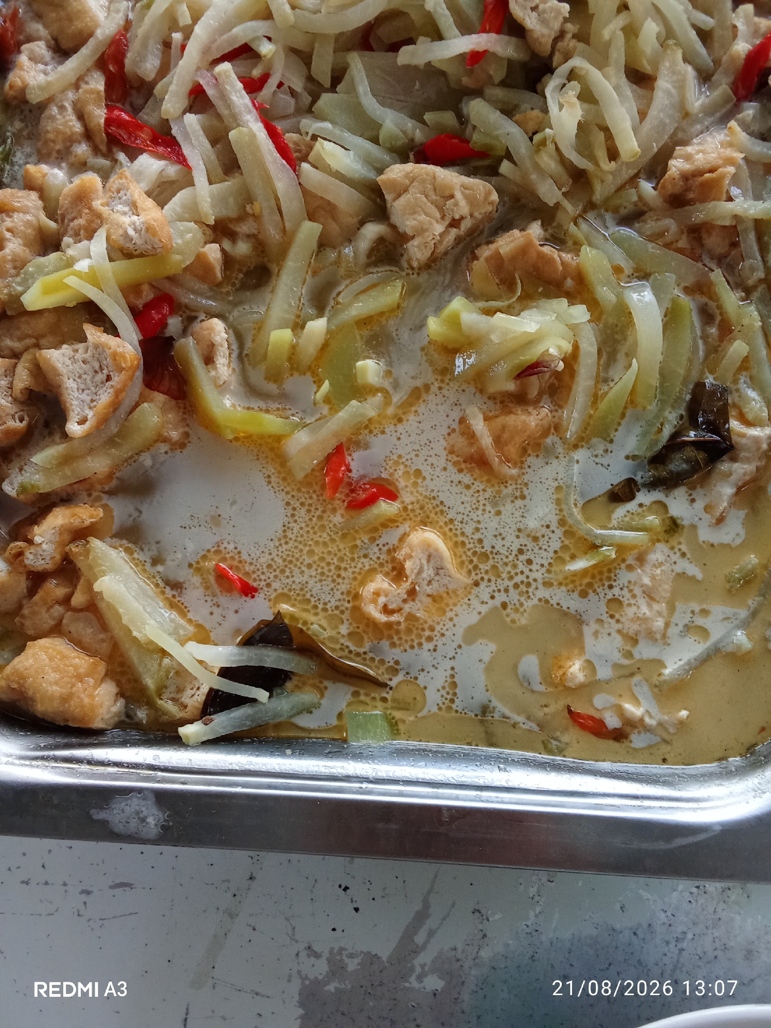 A close-up, overhead shot shows a metal tray filled with a dish of sliced chayote and tofu in a creamy, yellowish curry sauce. Red chili slices are scattered throughout the dish, adding color and likely spice. The tofu pieces are cubed and appear to be fried. The sauce has a slightly oily sheen with small bubbles, suggesting it has been recently cooked or is being kept warm. The metal tray itself is clean but shows some wear. The background is out of focus, but appears to be a slightly textured, possibly tiled surface. The image is sharp and well-lit, highlighting the textures and colors of the food. The date stamp in the lower right corner indicates the image was taken on August 21, 2026, at 13:07. There are no people or other identifiable objects visible that would place the scene in Banaran, Indonesia.